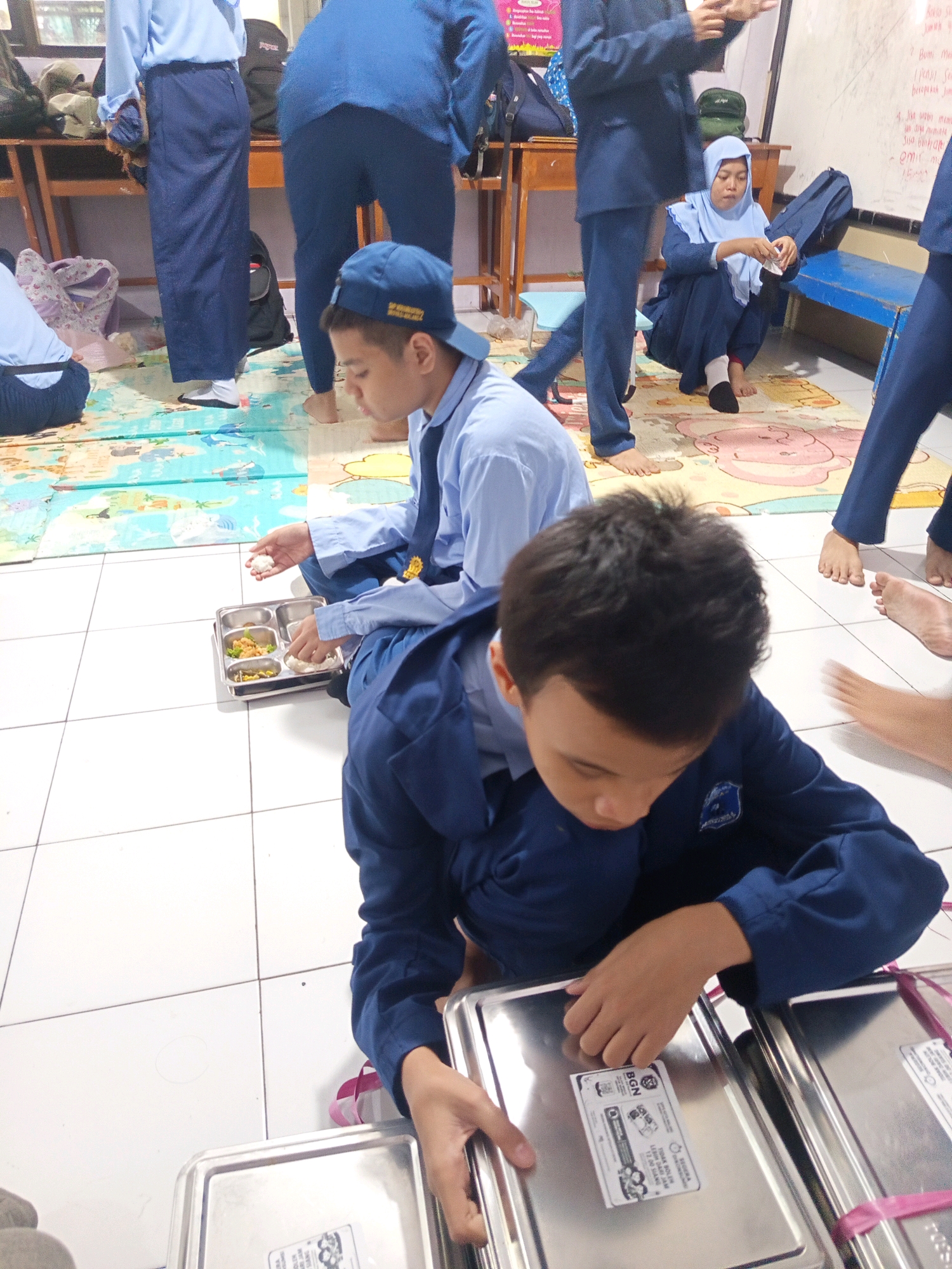 In a classroom or communal space, likely in Malang, Indonesia, several students in blue uniforms are seen sitting on a mat. The mat is colorful and features cartoonish pink figures. The floor is tiled with white squares.
In the foreground, a young student wearing a blue uniform and a blue cap is looking down at a metal lunch tray. The tray contains several compartments with food, including rice. The student is holding a piece of rice in their hand. To the right of this student, another student's hands are visible, reaching for or interacting with a stacked metal food container, tied with pink ribbon.
In the background, another student in a blue uniform is seated on the mat, looking towards the food tray. Further back, a student wearing a light blue hijab and a blue uniform is also seated on the mat, holding something in their hands and looking down. Several blue backpacks and other bags are visible, as well as wooden furniture. The overall scene suggests a mealtime or distribution of food within a school setting.