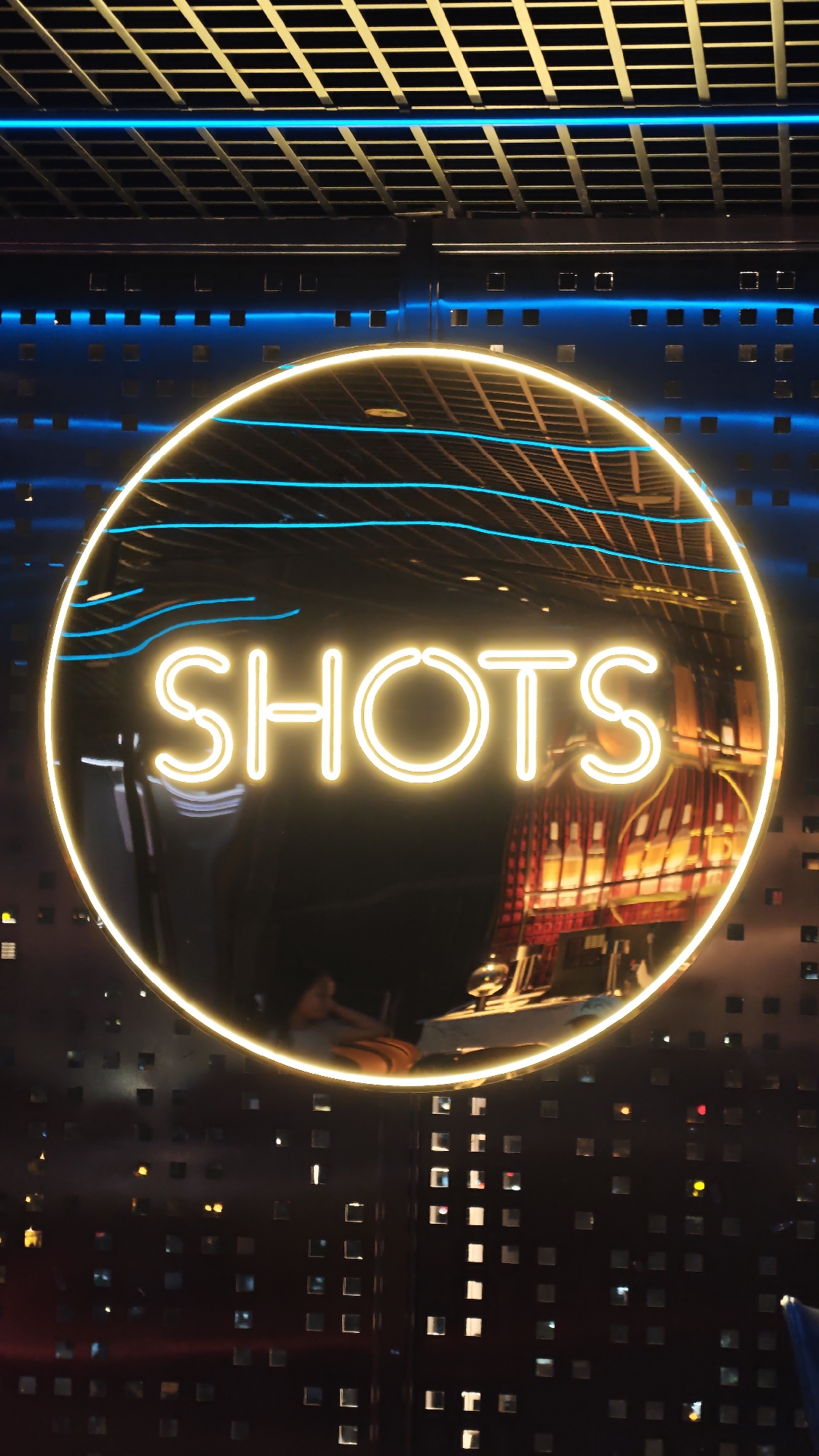 The image displays an interior view of what appears to be a modern bar or entertainment venue, likely in Đà Nẵng, Vietnam, at night or in a dimly lit setting. The central subject is a large, circular neon sign, glowing brightly in yellow-orange, which prominently spells out the word "SHOTS."
The reflective surface of the circular sign provides a distorted but discernible view of the environment behind the viewer. Reflections show various objects, including multiple liquor bottles arranged on shelves, suggesting a well-stocked bar setup. A faint, indistinct reflection of a person, possibly seated or leaning, is also visible within the lower part of the circle.
In the background, above the main sign, a ceiling structure with a grid pattern, possibly metal, is illuminated with a warm light. Below this ceiling grid, several horizontal blue neon lines or strips are visible, adding to the venue's vibrant lighting scheme. The walls surrounding the circular sign are dark and textured, featuring numerous small, square cutouts. Many of these squares are lit from within or behind, creating a pattern of glowing points that contribute to a futuristic or stylish aesthetic. Additional blue neon accents are scattered across the background, enhancing the dynamic atmosphere.
The visible text "SHOTS" directly indicates the type of establishment, reinforcing the impression of a lively drinking spot. The overall scene suggests a contemporary, energetic environment designed for social gatherings.