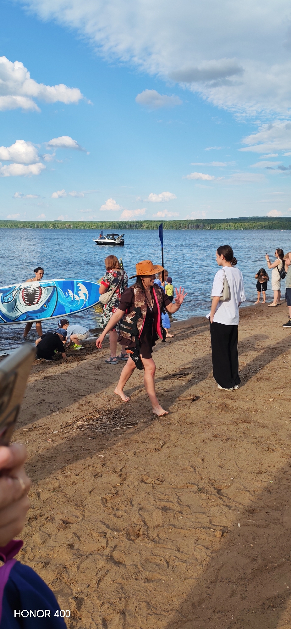 The image depicts a sunny day at a beach on the Kama River in Chaikovsky, Russia. The scene is populated with several individuals engaged in various activities near the water's edge.
In the foreground, a person wearing a brown cowboy hat and a camouflage vest is walking barefoot on the sandy shore, waving with their right hand. They are dressed in a dark dress and have a pink flotation device around their waist. To the left of this person, a woman in a floral dress is standing near a paddleboard decorated with a shark design. Children are visible in the shallow water, with one child digging in the sand and another sitting near them.
Further back, a group of people are gathered near the water, including a woman in a white t-shirt and black pants, who appears to be facing away from the camera, and another woman holding a phone, possibly taking pictures. A white boat with a black top is visible in the middle ground on the blue water, and a line of trees forms the background along the riverbank. The sky is blue with scattered white and gray clouds. The sand on the beach shows numerous footprints and some scattered dry twigs. The overall atmosphere suggests a casual outdoor gathering by the river.