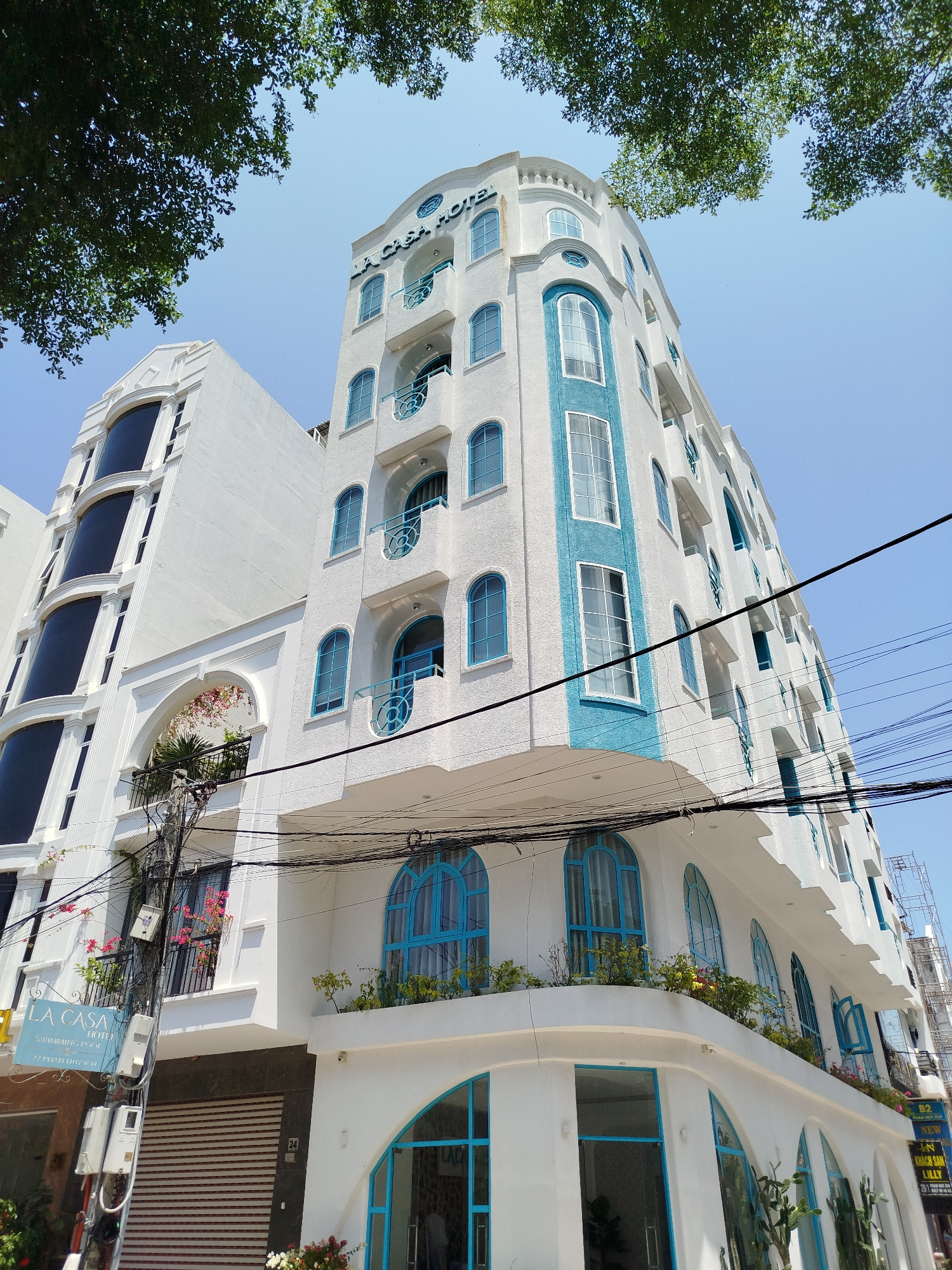 The image depicts a view of the La Casa Hotel in Ho Chi Minh City, Vietnam, as indicated by the signage present. The hotel is characterized by its white facade with blue accents on the windows and balconies. The architectural style incorporates arched windows and decorative railings. Power lines are visible in the foreground, crossing the view of the buildings. Additional signage and another building are seen adjacent to the hotel, indicating its location within an urban environment. Trees are seen at the top and bottom of the image.
