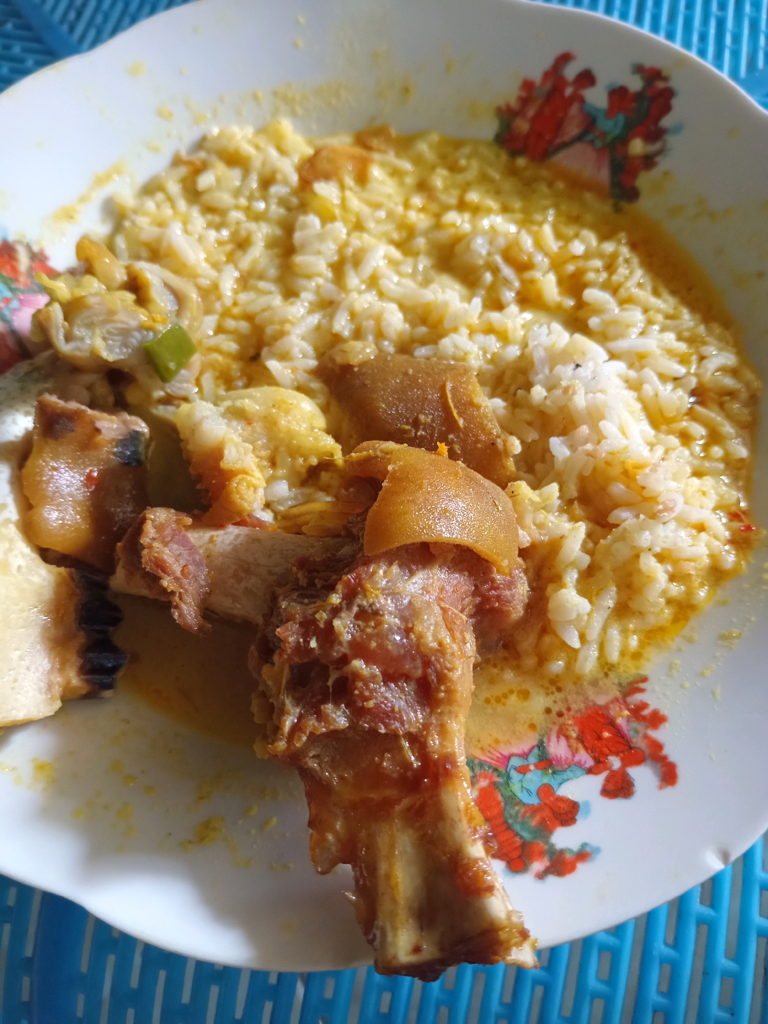 A close-up, overhead shot showcases a white plate with a colorful floral design on its rim, holding a portion of rice and meat. The rice, appearing somewhat mushy and mixed with a yellow sauce, is piled in the upper right quadrant of the plate. Mixed with the rice are what appear to be small pieces of scrambled egg or curdled sauce. Dominating the left and lower portions of the plate are several pieces of meat, some with bone and skin. One large piece of bone with meat attached sits prominently in the foreground, with other smaller pieces scattered around it. The overall dish appears to be a hearty meal. The plate rests on a blue woven placemat. The scene is likely a kitchen or dining area in Randuagung, Indonesia, given the context provided.