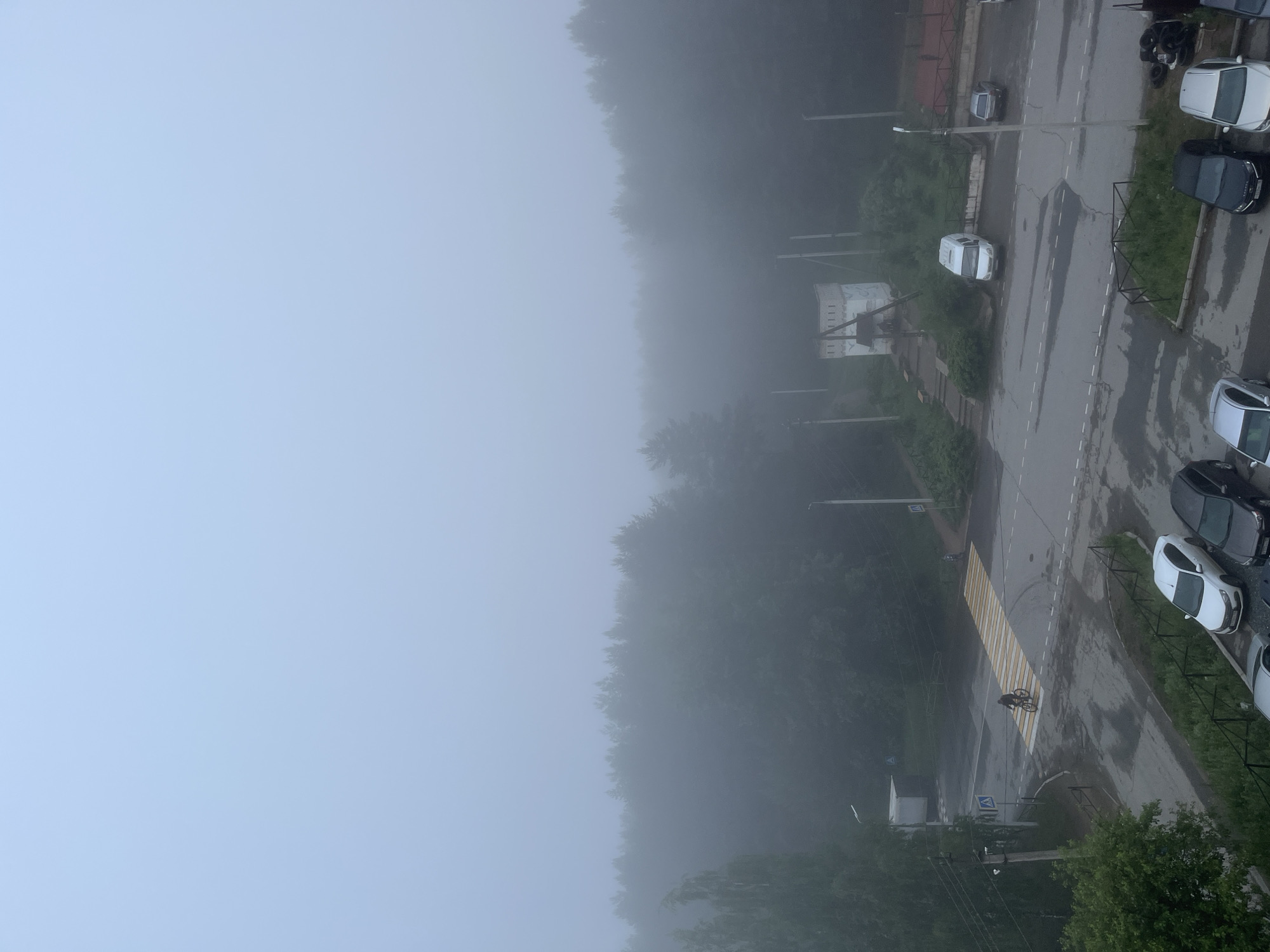 The image presents a high-angle view of a street in Chaikovsky, Russia, shrouded in dense fog. The pervasive mist significantly limits visibility, creating a subdued atmosphere.
The upper-left portion of the frame is dominated by a pale, featureless sky, heavily obscured by the fog. A thick band of dark green foliage, indicative of a forest or dense woodland, stretches horizontally across the mid-ground, its details softened by the atmospheric conditions.
In the lower-right section, a wet asphalt road is visible, curving slightly. A prominent pedestrian crosswalk with alternating yellow and white stripes diagonally traverses the road. A solitary person on a bicycle is captured mid-crossing on these stripes. Numerous vehicles are parked along the edges of the road, including cars of white, black, and silver hues. Another white van-like vehicle is seen on the road itself. Streetlights or utility poles are intermittently spaced along the roadside. Near some parked cars on the far right, a small pile of tires or industrial debris is noticeable. Faint outlines of a distant building can be glimpsed through the fog within the tree line.
The diffused lighting suggests either an early morning or late afternoon setting, consistent with the heavy fog. The ground's wet appearance points to recent rain or high humidity. No legible text is visible on any objects, signs, or vehicles.