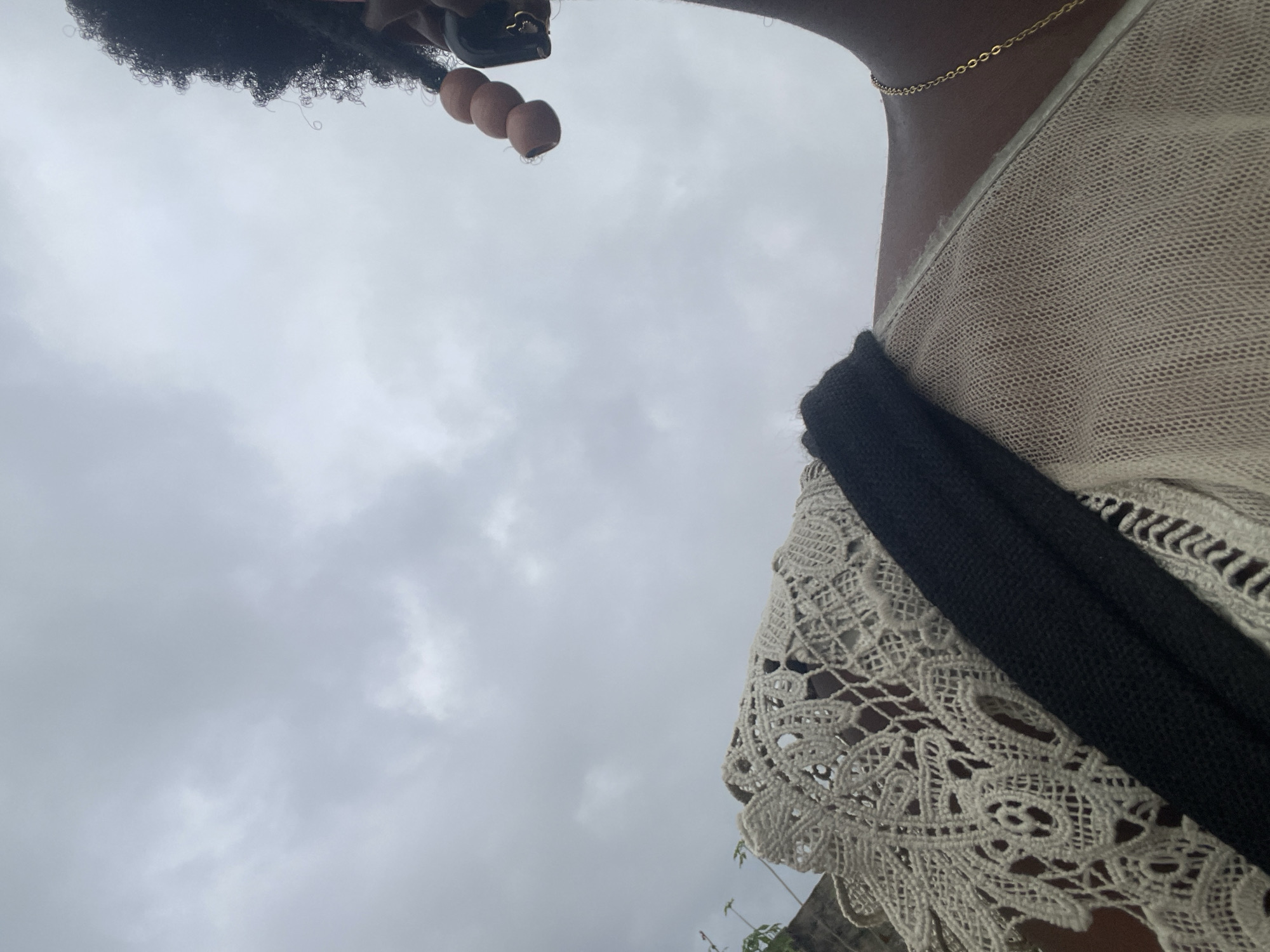 A close-up, low-angle shot features a person's shoulder and head against an overcast sky. The person is wearing a beige knitted top with a black strap and a white lace collar. Their hair is dark and curly, adorned with light brown beads. A thin gold chain necklace is visible on their neck. The background is a cloudy, gray sky. The scene is located in Iyafin, Nigeria.