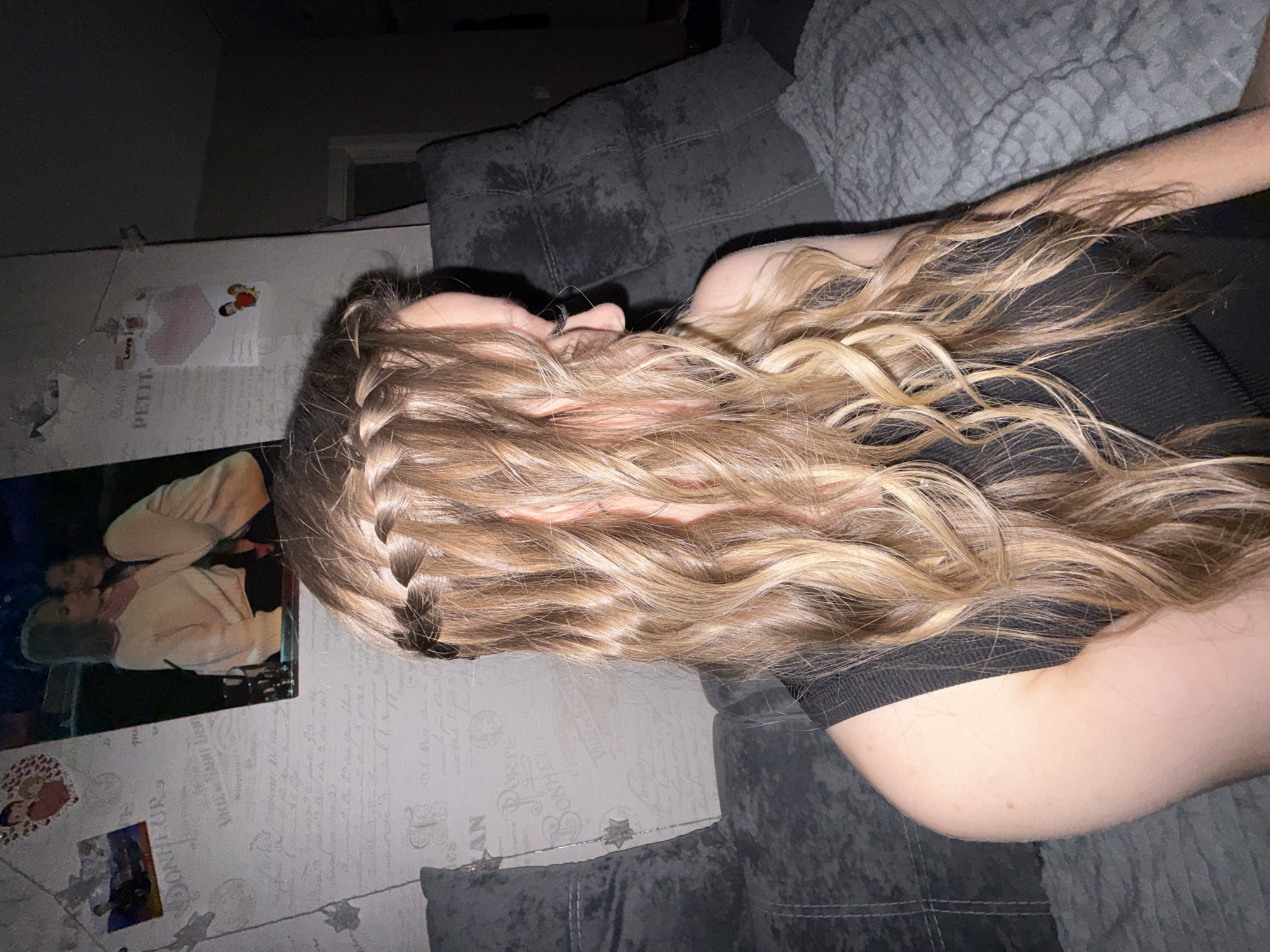 A young woman with long, wavy blonde hair is shown from the chest up, from a side profile. Her hair is styled with a braid that runs across the top of her head, with strands of hair falling out to create a waterfall effect. She is wearing a black, ribbed top. The background shows a textured wall with framed pictures and a dark gray sofa. The scene appears to be indoors, possibly a living room. There is no indication of the city of Chaikovsky, Russia.