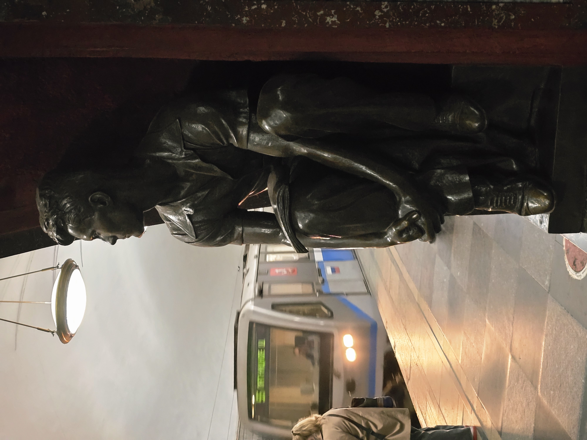 The image shows a bronze statue of a man in a squatting position at the Stockholm Metro Station in Stockholm, Sweden. A metro train is passing behind the statue. A person is waiting for the metro on the platform. Overhead lighting is present.