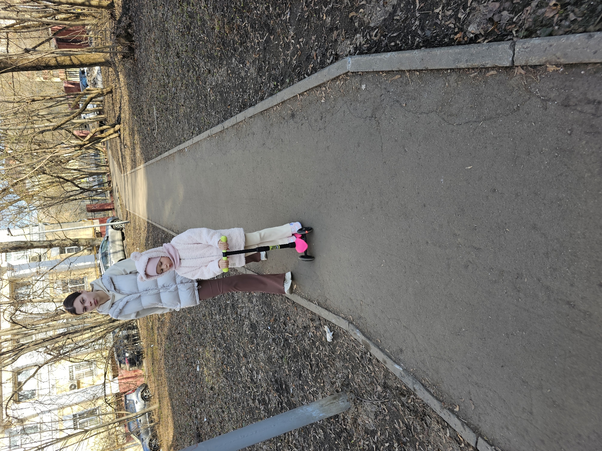 A woman and a young child on a scooter are walking on a paved path in Moscow, Russia. The path is lined with bare trees and buildings in the background. The woman is wearing a light-colored puffer jacket and brown pants. The child is wearing a pink jacket with a hood and riding a pink scooter with green handles.