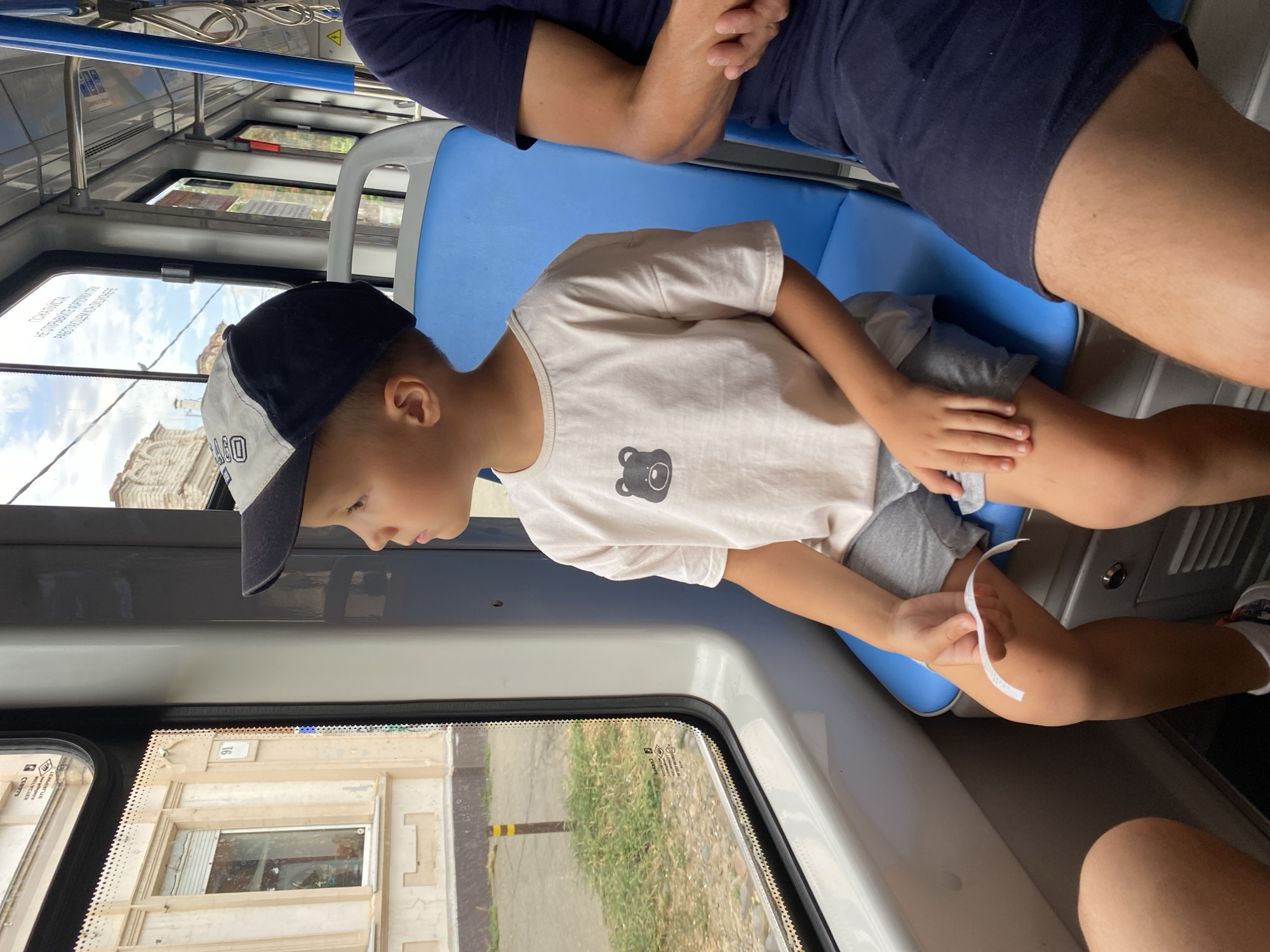 In this image, a young boy wearing a black and white baseball cap and a white t-shirt with a bear graphic is seated on a blue bus seat. He is looking out of the window, with his head slightly tilted. His hand is holding a small white piece of paper. To his left, another person's legs are visible, wearing dark blue shorts and a dark blue shirt. The boy is in a bus, with the interior of the bus visible, including metal poles and windows. Outside the window, buildings and trees can be seen, suggesting an urban or semi-urban environment. The lighting suggests it is daytime, likely sunny. The image appears to have been taken from inside the bus, looking towards the boy. The overall mood is casual and observational.