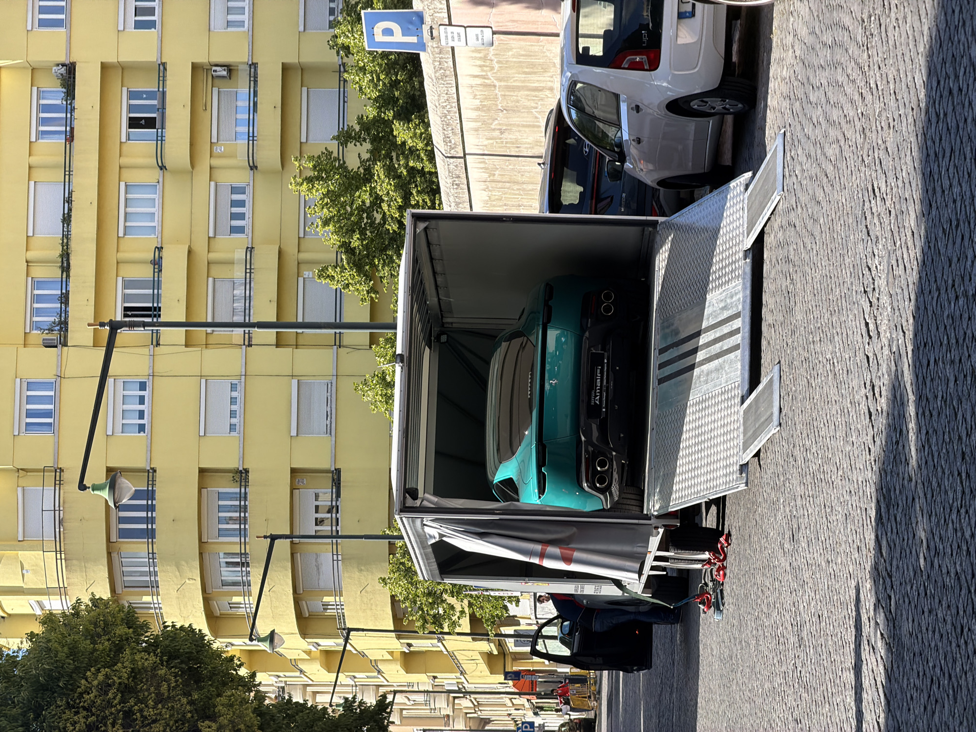 A turquoise sports car is secured inside a silver trailer parked on a cobblestone street in Lisbon, Portugal. The car's rear is visible, featuring four exhaust pipes and the words "Ferrari Amalfi." The trailer is open at the back, revealing the car inside. In the background is a yellow apartment building with many windows. A parking sign is visible near the front of the trailer. A black car with an open door is parked near the trailer's front end. The sky is clear, indicating daylight.