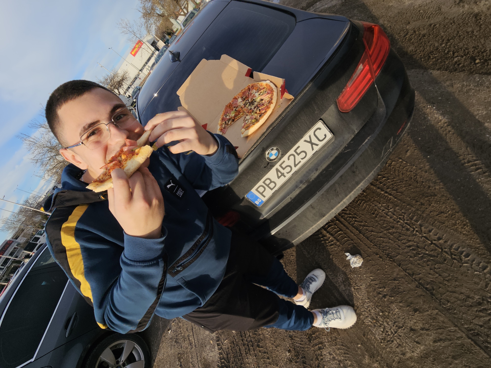 A young man with short dark hair and glasses is eating a slice of pizza in a parking lot. He's wearing a blue and yellow Puma hoodie and black pants, and white sneakers. A pizza box containing the remaining portion of the pizza rests on the trunk of a black BMW. The car's license plate reads "PB 4525 XC," which indicates that the car is registered in Bulgaria. The parking lot surface is dirt and has tire tracks. In the background, there is a building with a "Kaufland" sign, which is a European hypermarket chain. Given the location, it is plausible that the image was taken in Plovdiv, Bulgaria, which has a Kaufland store. A crumpled tissue or paper is on the ground near the man's feet.