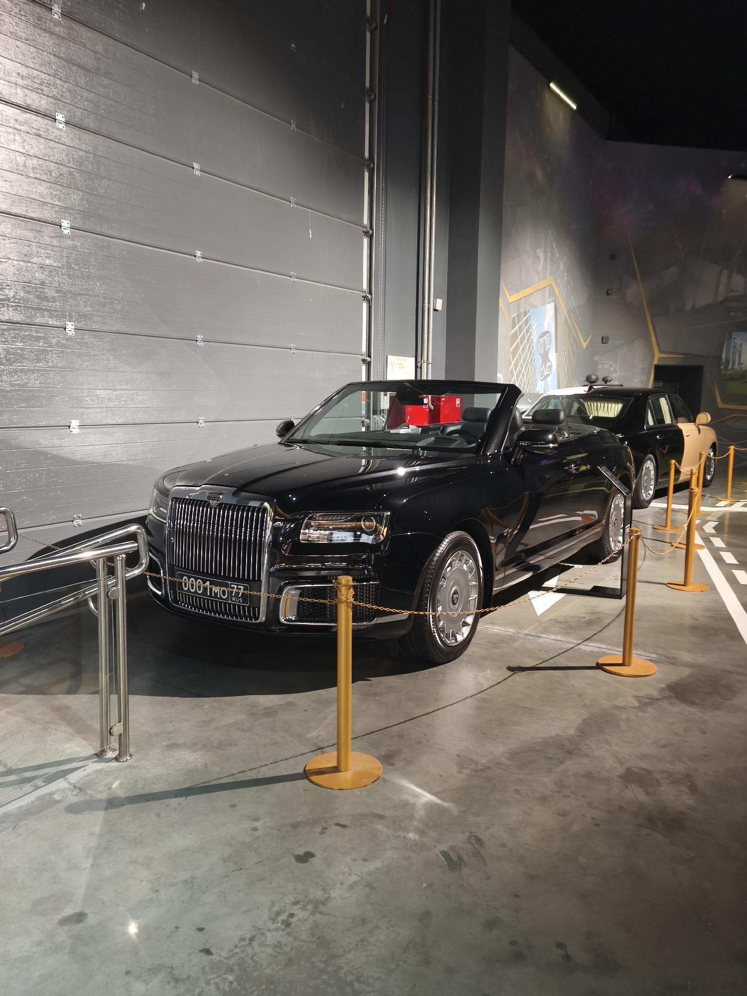 The image displays a black convertible luxury car, possibly a prototype or custom build, parked indoors. The car has a chrome grille, distinctive headlights, and custom wheels. A license plate with "0001 MO 77 RUS" is visible on the front. In the background, another black and tan car is parked. The environment appears to be a showroom or exhibition space, indicated by the polished concrete floor, metallic grey paneling on the wall, and stanchions with yellow chains to cordon off the vehicle. The wall behind the cars features a mural depicting a cityscape and perhaps a space-themed design. The lighting is artificial and dramatic, casting shadows on the floor. This scene is likely from a car exhibition or a private collection, and given the "77" on the license plate, which typically denotes Moscow in Russian vehicle registration, the location is in the city of Moscow, Russia. There are no people visible in the image.