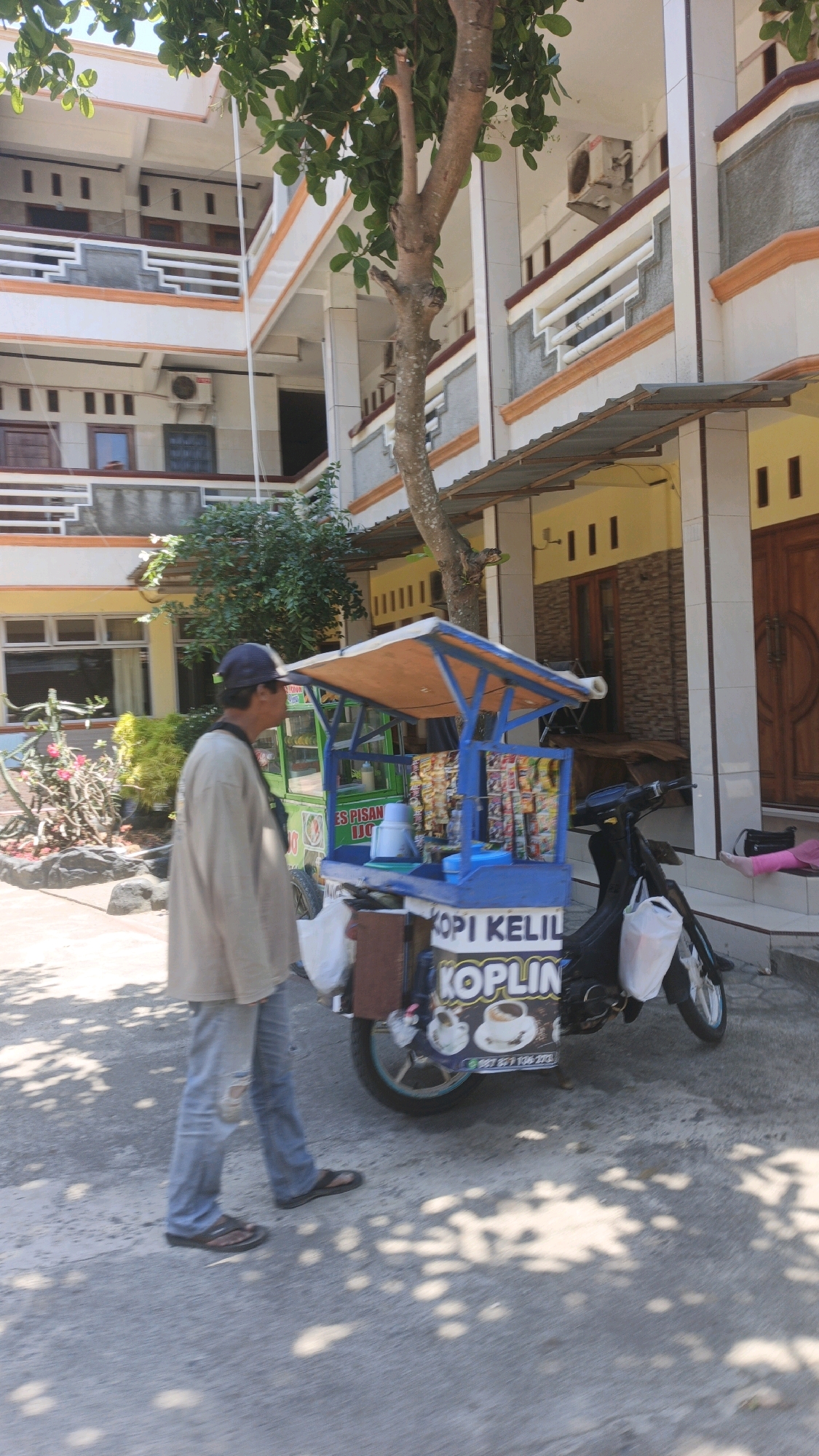 A man in a light-colored shirt and blue jeans walks past a food cart in front of a multi-story building in Desa Pagandaran, Indonesia. The food cart, which is mounted on a motorcycle, is blue and yellow and displays various snacks and drinks. The building appears to be a residential or commercial building with multiple windows and balconies. The street is paved and has some shadows cast by trees. The man is walking away from the camera, and his face is not clearly visible.