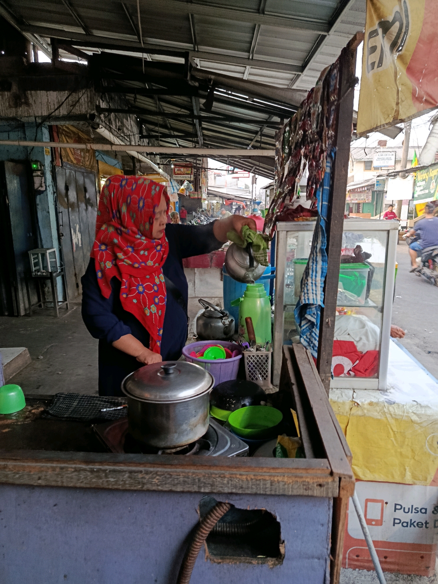A woman wearing a red hijab with a floral pattern and a dark blue top is standing behind a food stall in a market in Bekasi, Indonesia. She is holding a metal kettle with a green cloth wrapped around its handle and appears to be pouring liquid into a green container. In front of her is a metal pot on a stove, a purple basket with green and pink bowls, and a basket with utensils. To her right is a glass display case containing various items, including red fabric. The stall is covered by a metal roof and has various signs and banners in the background. People and motorcycles are visible in the blurred background, indicating a busy street or market environment. The overall scene depicts a vendor at work in an outdoor marketplace.