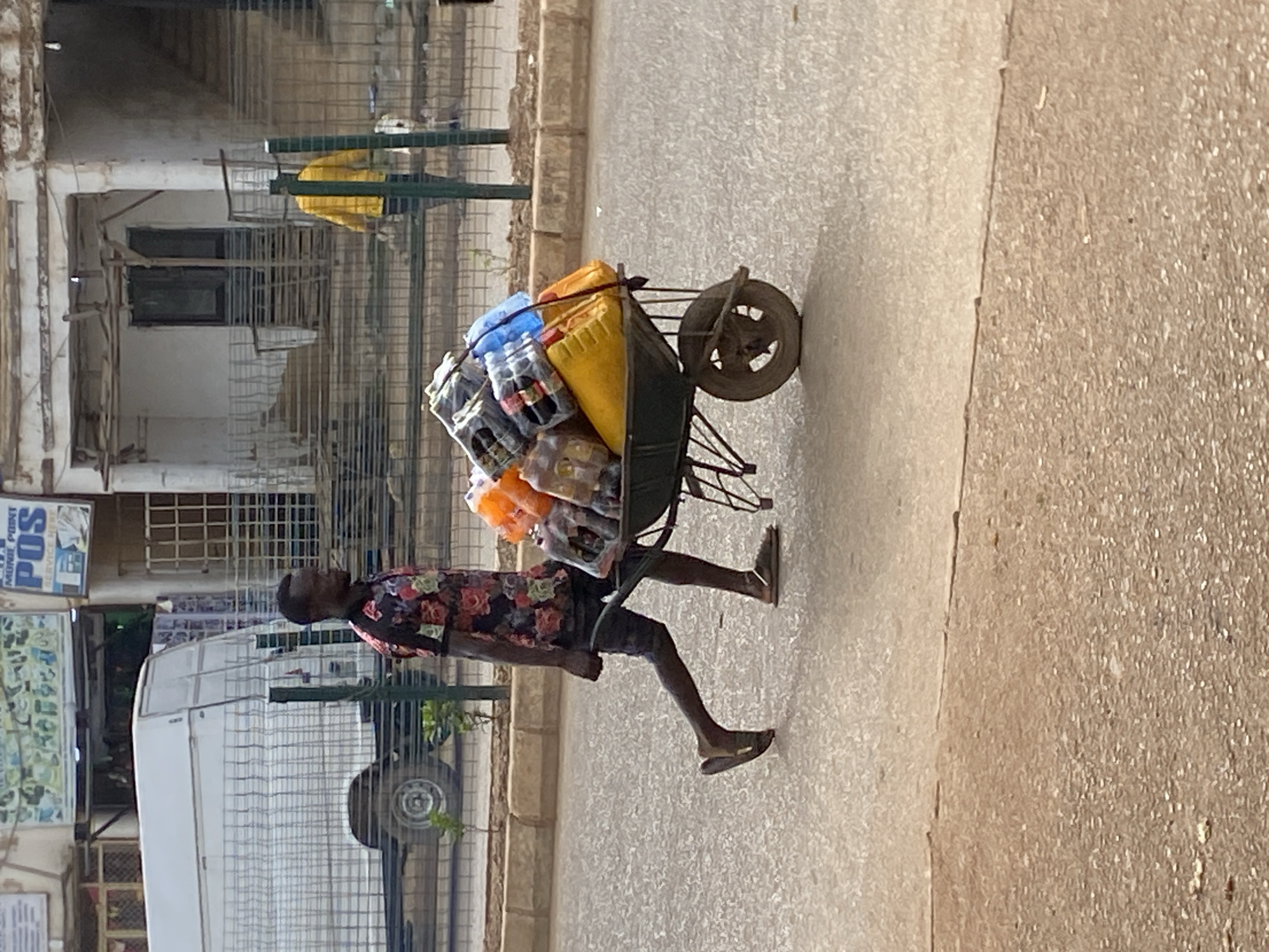 A person is pushing a wheelbarrow loaded with plastic containers, likely for water, down a paved street in Bukuru, Nigeria. The person, who appears to be male with dark skin, is wearing a colorful floral shirt and dark shorts. He is also wearing sandals. The wheelbarrow is dark green and has a single wheel. The containers are of various colors, including yellow, blue, and orange, and some appear to be filled with liquid. Some of the containers are stacked and secured with ropes.
The scene is outdoors, with buildings and a fence visible in the background. One building has barred windows. There's a white vehicle with a metal cage on top parked to the left of the frame. Another sign with blue text is visible on the building in the background, with the word "POS" clearly legible. The ground is paved, and the lighting suggests it is daytime, likely late morning or early afternoon, with bright sunlight casting shadows. The weather appears to be clear and sunny. The overall impression is of a busy street scene with a person engaged in transporting goods.