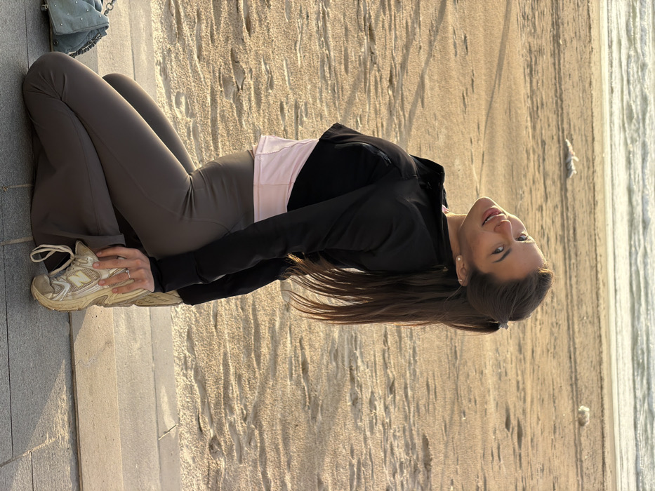 A female subject is depicted kneeling on a dark gray tiled promenade adjacent to a sandy beach in Tel-Aviv, Israel, during golden hour lighting. The Mediterranean Sea is visible in the background with gentle waves. The subject, with long dark hair tied in a ponytail, wears a light pink top, a black cropped jacket, dark gray leggings, and white and beige athletic sneakers. She is smiling directly at the camera with her head tilted back and to her left. A ring with a clear stone is on her left ring finger, and a light blue quilted handbag is on the pavement beside her. Footprints and two indistinct light-colored objects are visible on the sun-kissed sand.