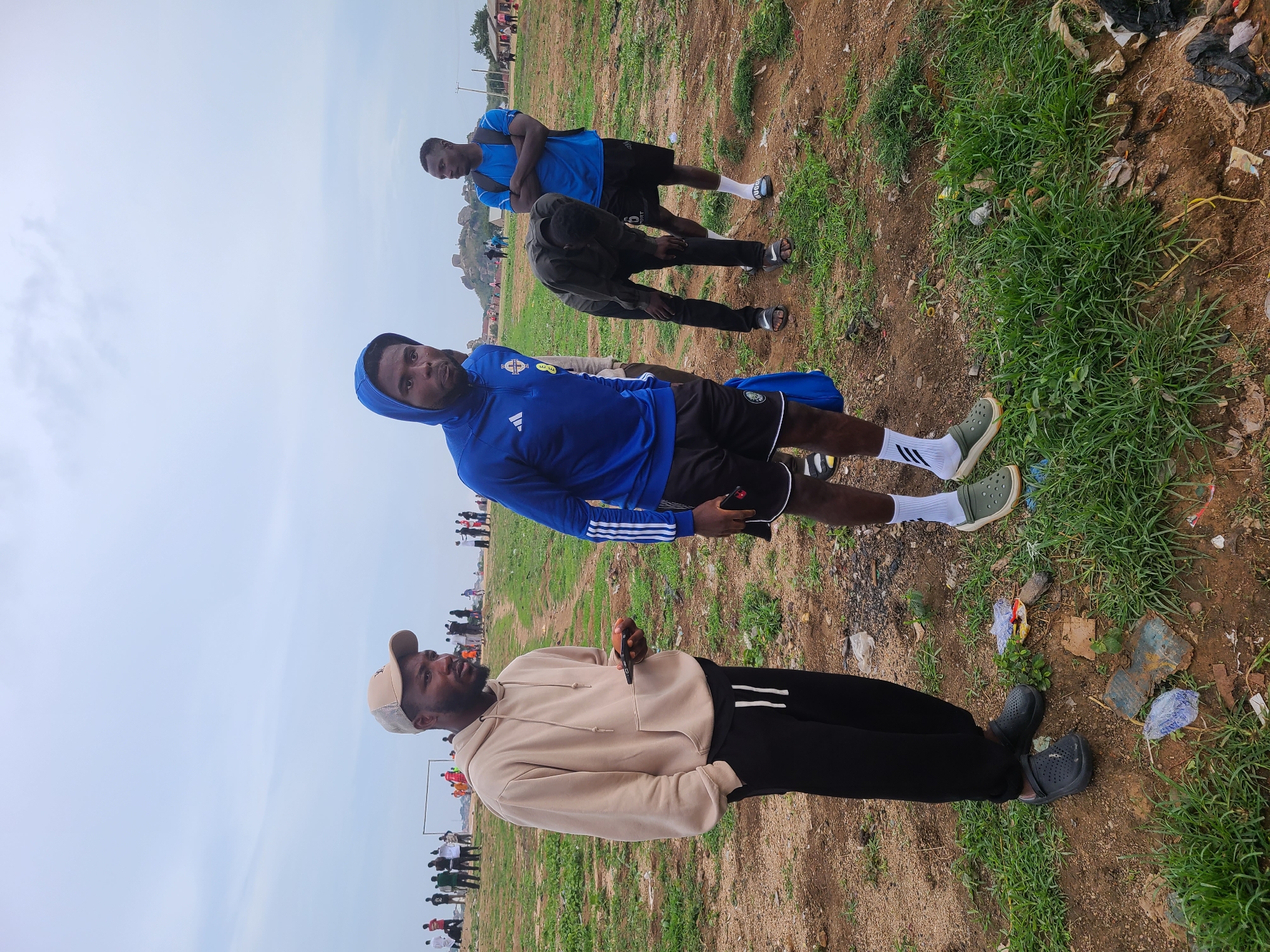 The image shows four men outdoors in a field in Unknown, Unknown. The man in the foreground wears a tan hooded sweatshirt, black sweatpants with two white stripes on the outer seams, a light-colored baseball cap, and black clogs. He holds a dark object in his right hand. Behind him stands a man in a blue hooded jacket with white stripes on the sleeves and black shorts, wearing white socks with dark stripes and olive green sneakers. Further back, two men in blue shirts and black shorts appear to be stretching. The ground is a mix of dirt and green grass. Debris and trash are scattered on the ground, particularly on the right side of the image. In the background, a crowd of people stands near a sports field with white goalposts. The sky is overcast.
