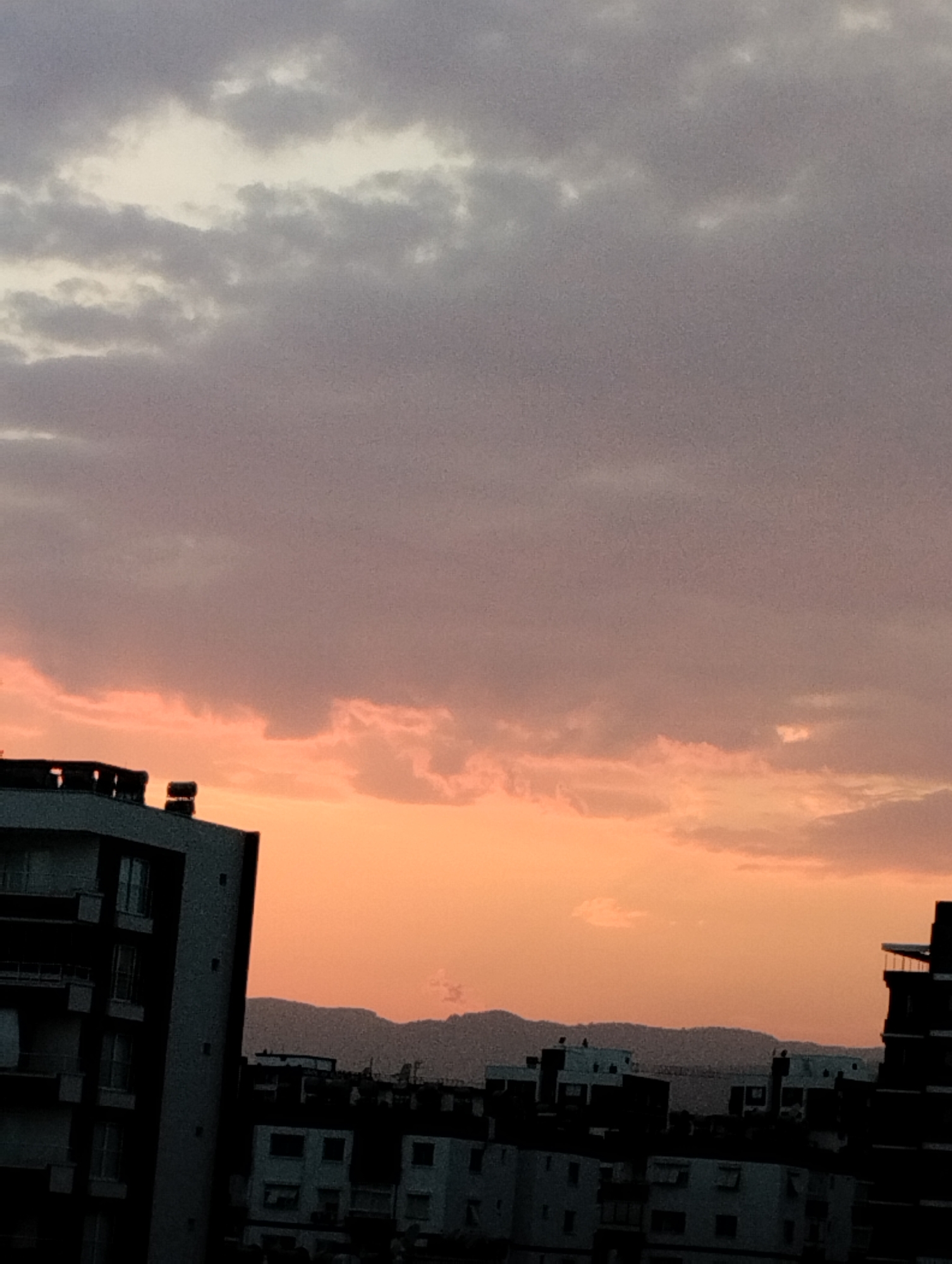 This image depicts a dusk scene over Mezitli, Turkey. The sky is dominated by clouds, with a gradient of colors ranging from deep gray at the top to a warm orange and pink hue near the horizon. The sun is not visible, suggesting it has recently set or is about to rise. Silhouetted against the colorful sky are the rooftops and upper floors of several multi-story buildings. In the foreground, on the left, a dark, modern building with multiple balconies is prominent. Further back, other buildings with visible windows and balconies are visible. In the distance, a range of low, indistinct mountains forms the horizon. The overall impression is one of a city settling into twilight.