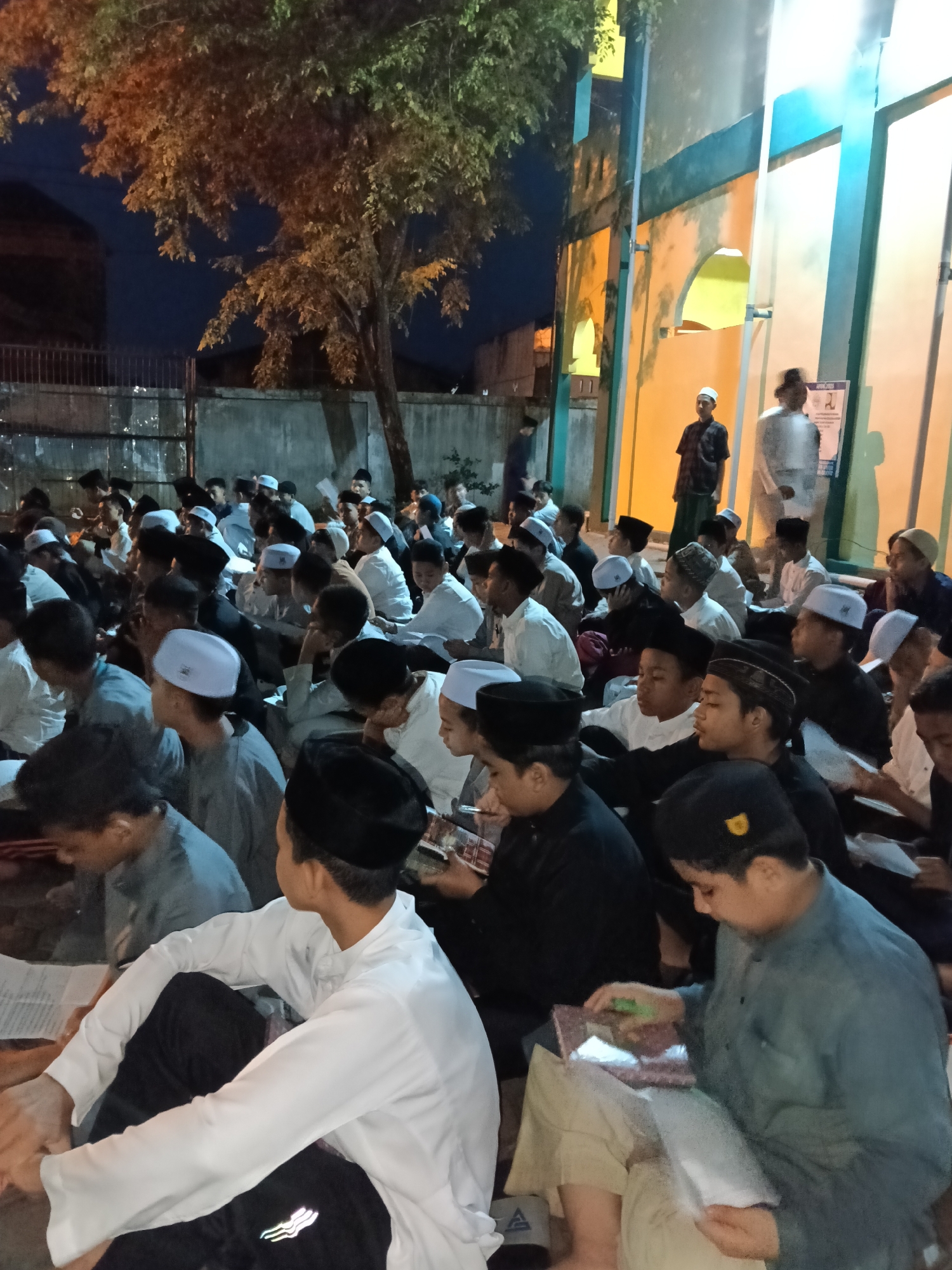 A large group of people, primarily young males, are sitting on the ground in an outdoor area in front of a building. The individuals are dressed in a mix of casual and religious attire. Many wear traditional Muslim caps (peci or kopiah) and some have white or light-colored shirts. Some appear to be reading from books. The location appears to be a mosque or religious center, possibly in Lhokseumawe, Indonesia, as this city is known for its large Muslim population. The building is painted in light colors with blue trim. The time of day appears to be evening or night, as it is dark and the building is lit with artificial lights. A tree is also visible near a fence in the background. Some of the people are interacting with each other, while others are focused on reading. The environment suggests a community event, possibly a religious gathering or study session.