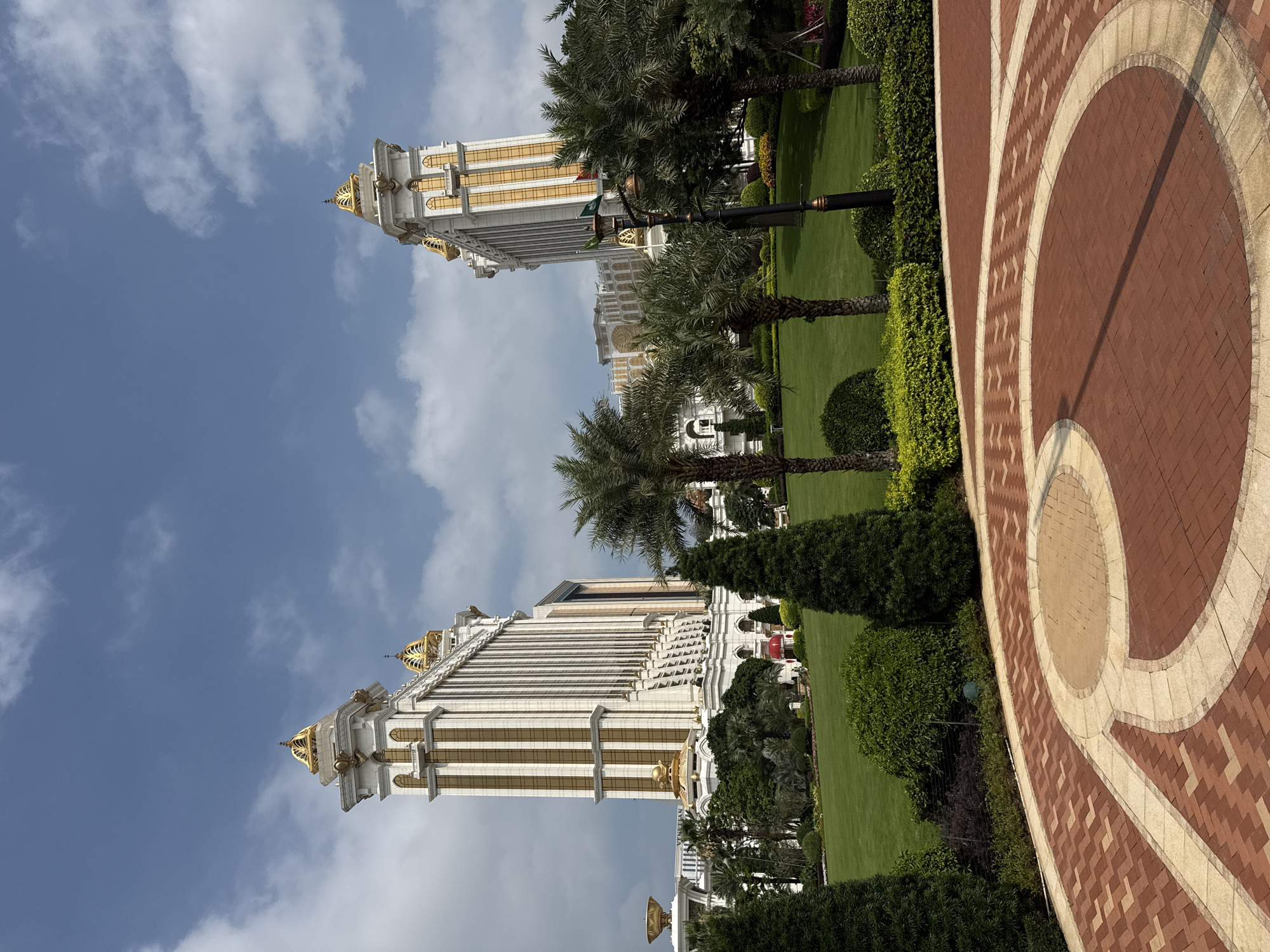 The image presents a view of a large, ornate building in what appears to be a landscaped garden. The building is tall, white, with gold trim, and has a distinct architectural style. Lush green lawns and trees surround the building, and a paved walkway with a circular design is prominent in the foreground. The sky is blue with scattered clouds. The location is identified as being in Macao, China. No people are visible in the image. The primary subjects are the building and its surrounding landscape. The scene appears to be taken during the daytime, under natural light. The overall impression is one of opulence and meticulous design.