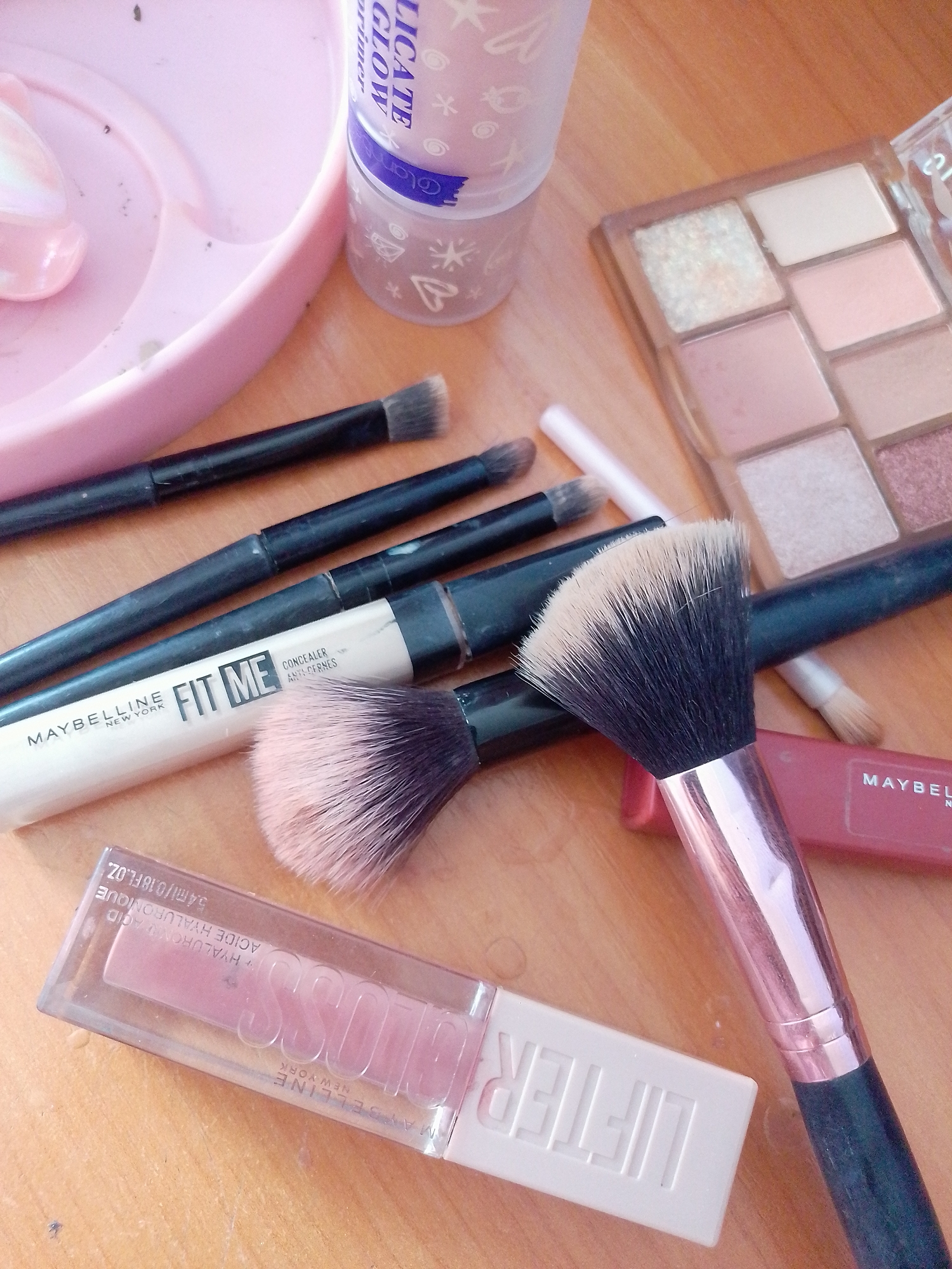 A flat lay of makeup products is arranged on a light brown wooden surface. In the upper left quadrant, a pink, heart-shaped tray contains a light pink, pearlescent item. To the right of the tray is a cylindrical container with a light purple lid, partially obscured, with white lettering that includes "GLOW" and "primer". A small eyeshadow palette with nine square pans of various neutral and pink shades is positioned in the upper right quadrant.
Several makeup brushes are scattered across the surface. Three black-handled brushes with grey bristles are visible, laid out horizontally. A concealer tube, labeled "FIT ME CONCEALER ANTI-CERNES" by Maybelline New York, lies beneath the brushes. A large, fan-shaped makeup brush with black and pink bristles and a metallic rose-gold handle is prominent in the center and right side of the image. Another smaller makeup brush with grey bristles is visible near the bottom right, partially obscured by the larger brush.
At the bottom of the image, a clear lip gloss tube with pink gloss inside is displayed horizontally. The tube is labeled with white lettering that includes "GLOSS" and "MAYBELLINE NEW YORK", and also "HYALURONIC ACID". A small pink lipstick or balm tube with "MAYBELLINE" written on it is partially visible beneath the fan brush.
The scene is domestic, likely a vanity or countertop. There are no people present. The lighting is bright, suggesting an indoor environment. The overall impression is one of personal grooming items. There is no information to place this scene within the city of Pryluky, Ukraine.