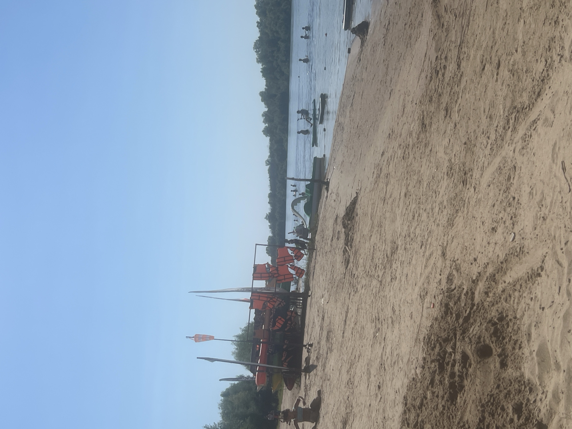 This image depicts a sunny day at a sandy beach along a wide river, likely the Volga River in Kilyakovka, Russia. The foreground is dominated by sand with some footprints and scattered debris. In the mid-ground, a rack holds several orange life vests and various watercraft, including kayaks and possibly a pedal boat. A young child is sitting on the sand in the left mid-ground. Further out on the river, several people are engaged in water activities, including paddling kayaks and swimming. The far bank of the river is lined with dense green trees under a clear blue sky.