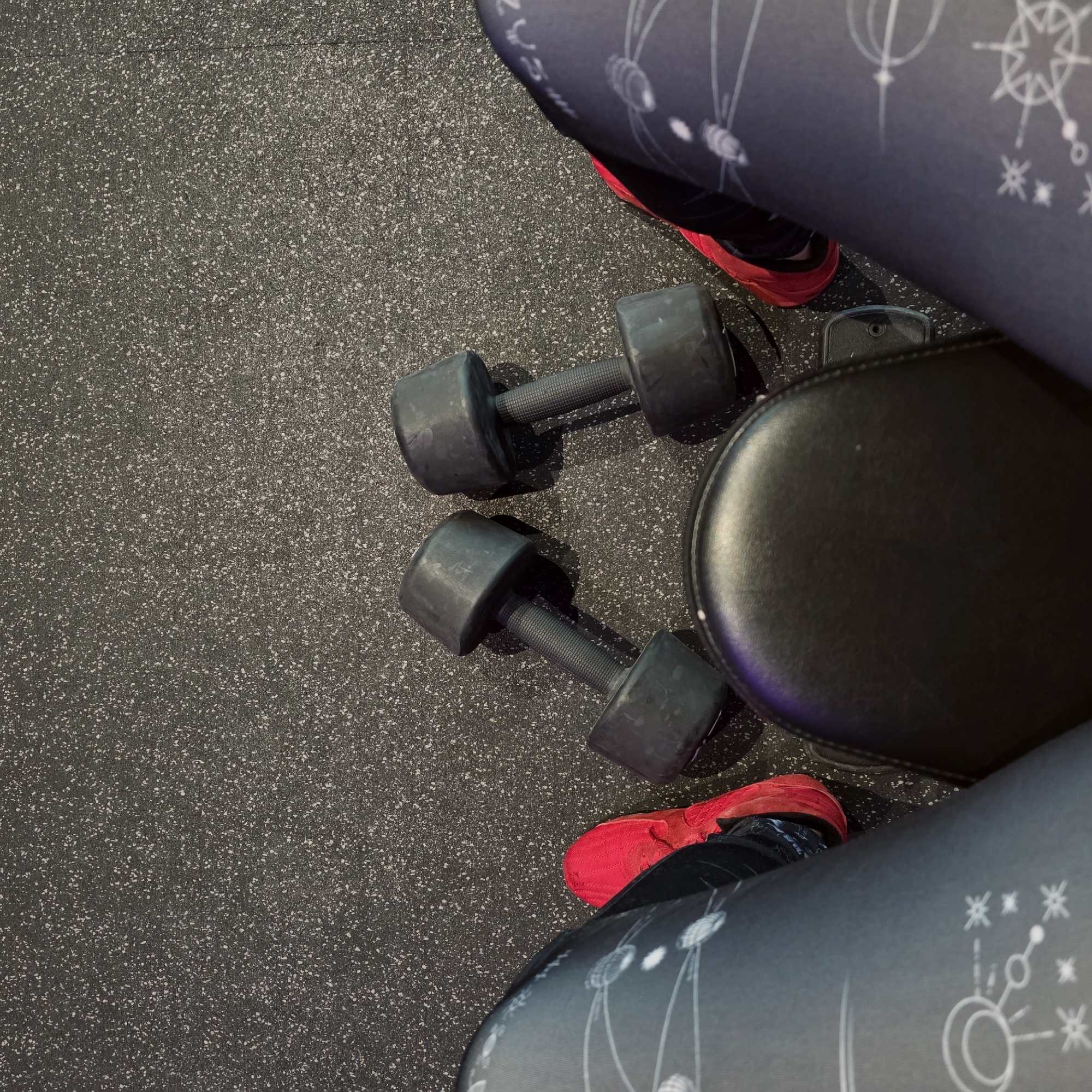 The image shows a close-up of a person at a gym. The person is wearing patterned leggings and red sneakers. Two black dumbbells lie on the speckled gray floor, along with a section of a black bench. There is no indication in the image that indicates that the location is in Saint Petersburg, Russia.