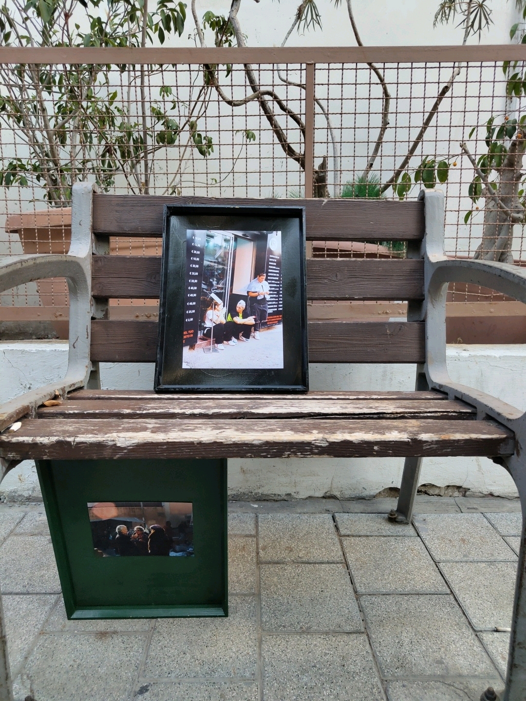 The image shows a weathered wooden park bench with a dark framed photograph resting on its backrest. The photograph appears to depict a group of people, possibly in an indoor setting, with text visible on the left side of the frame. In front of the bench, on the ground, is a dark green rectangular structure. Affixed to the front of this structure is another photograph, this one showing three people in what seems to be an outdoor, dimly lit environment. The bench is situated on a paved ground surface made of rectangular stone tiles. Behind the bench, a metal fence with a diamond pattern encloses greenery. The overall scene suggests an outdoor public space, possibly a park or a walkway, in Tel-Aviv, Israel.