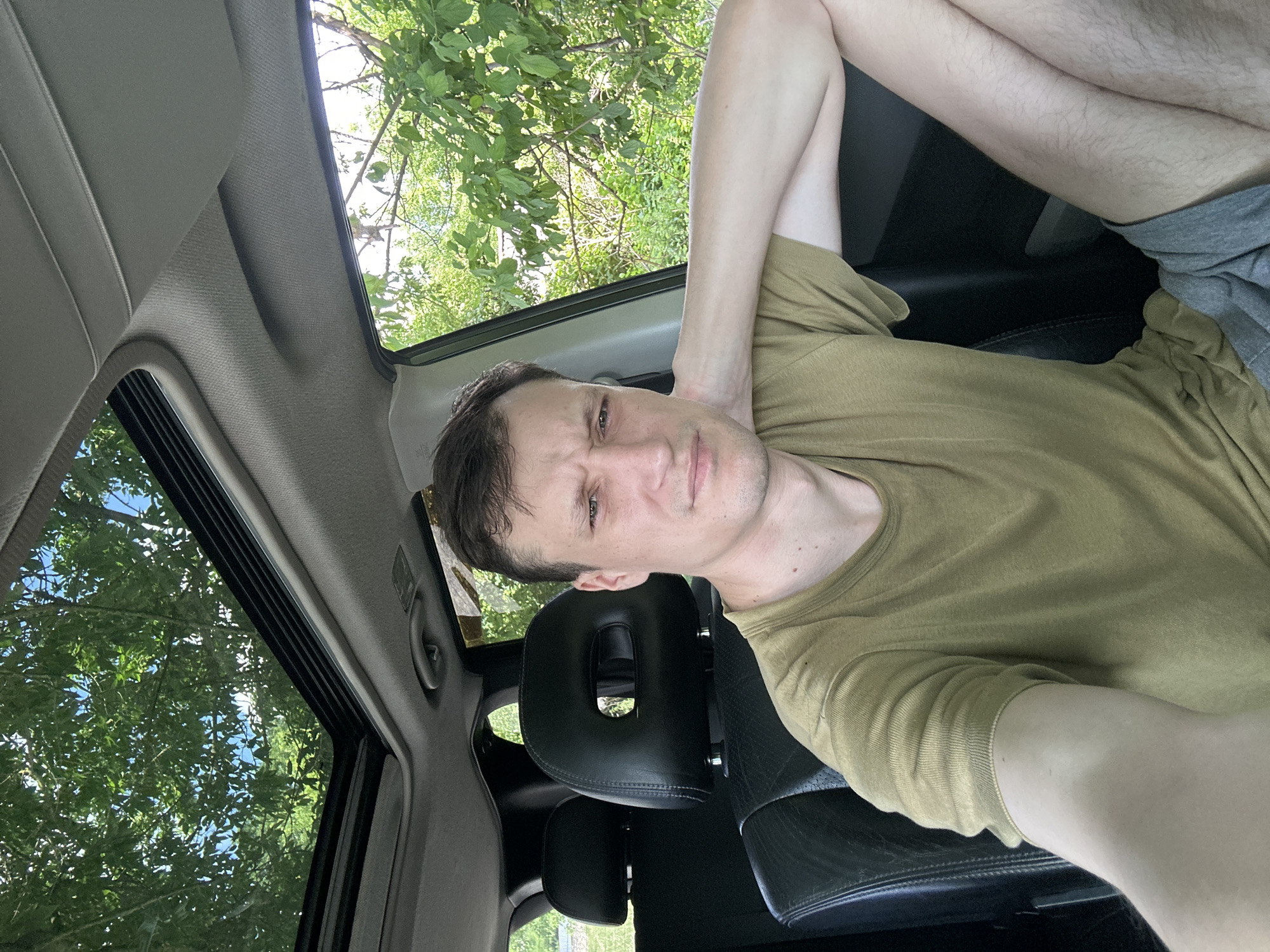 A man with short brown hair is sitting in the passenger seat of a car, looking at the camera with a displeased expression. He is wearing a light olive green t-shirt and grey shorts. His left arm is resting on the back of his head, and his right arm is bent at the elbow, with his forearm resting on his leg. The car has a sunroof open, revealing green trees and a blue sky. The interior of the car is visible, including black leather seats and headrests. The man appears to be in Makiv, Ukraine, based on the contextual information provided.