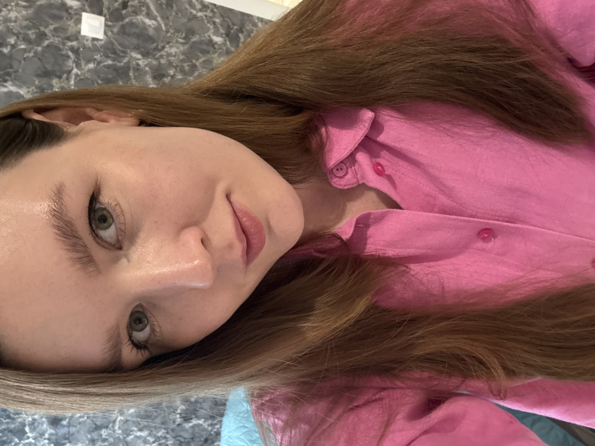 A young woman with light skin, hazel eyes, and light brown hair is seen from a close-up, slightly angled perspective. She is wearing a pink button-down shirt. The background features a gray patterned surface and a small, white rectangular object. The location is not discernible from the image.