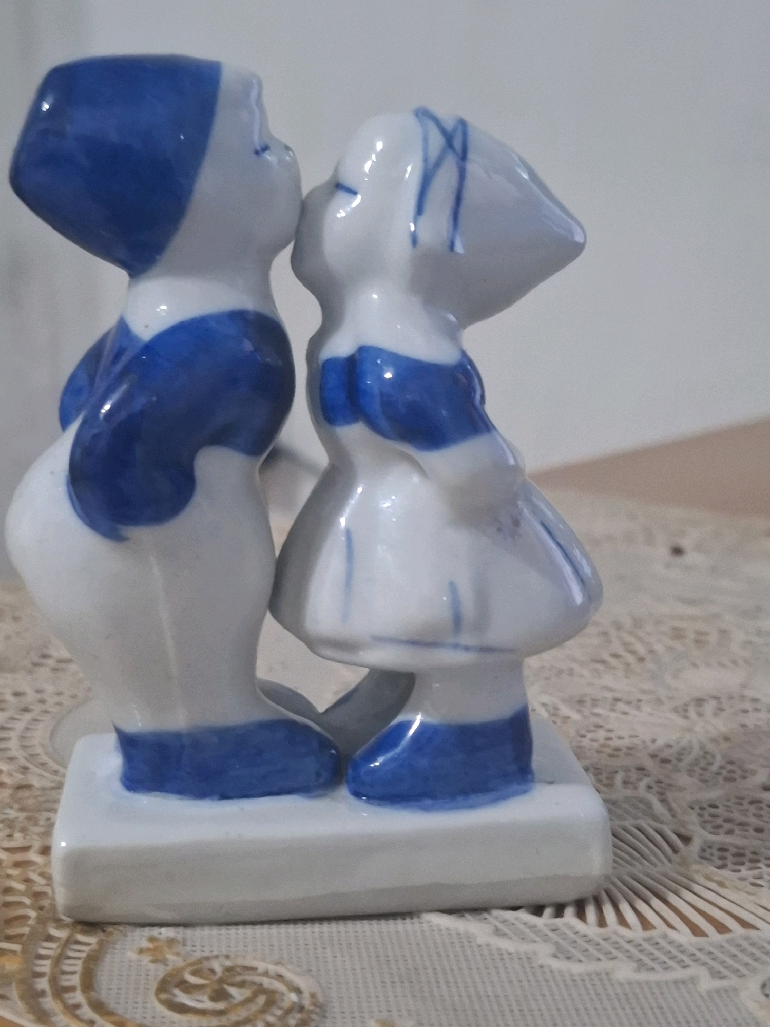 This is a close-up, eye-level, vertical shot of a decorative ceramic figurine featuring two young figures, a boy and a girl, in a romantic embrace. They are depicted in profile, leaning in for a kiss. The figurine is predominantly white with prominent blue detailing.
The boy, on the left, wears a blue cap that covers his hair and a blue top with white accents. His body is turned slightly away from the viewer, showing his rounded back. The girl, on the right, has blue lines drawn on her white cap, resembling hair. She wears a white dress with a blue collar and trim around the skirt. Her body is angled towards the boy, with her face close to his.
Both figures are standing on a white rectangular base. The figurine is placed on a textured surface, possibly a tablecloth or placemat, with a delicate, lace-like pattern in light brown and white. Behind the figurine, the background is a plain, light-colored wall, creating a simple backdrop that highlights the ceramic figures.
The scene captures a tender moment between the two figures, conveying innocence and affection. The lighting is soft and diffused, suggesting an indoor setting, possibly during daylight hours. There is no visible text in the image. The setting appears to be a domestic environment.
