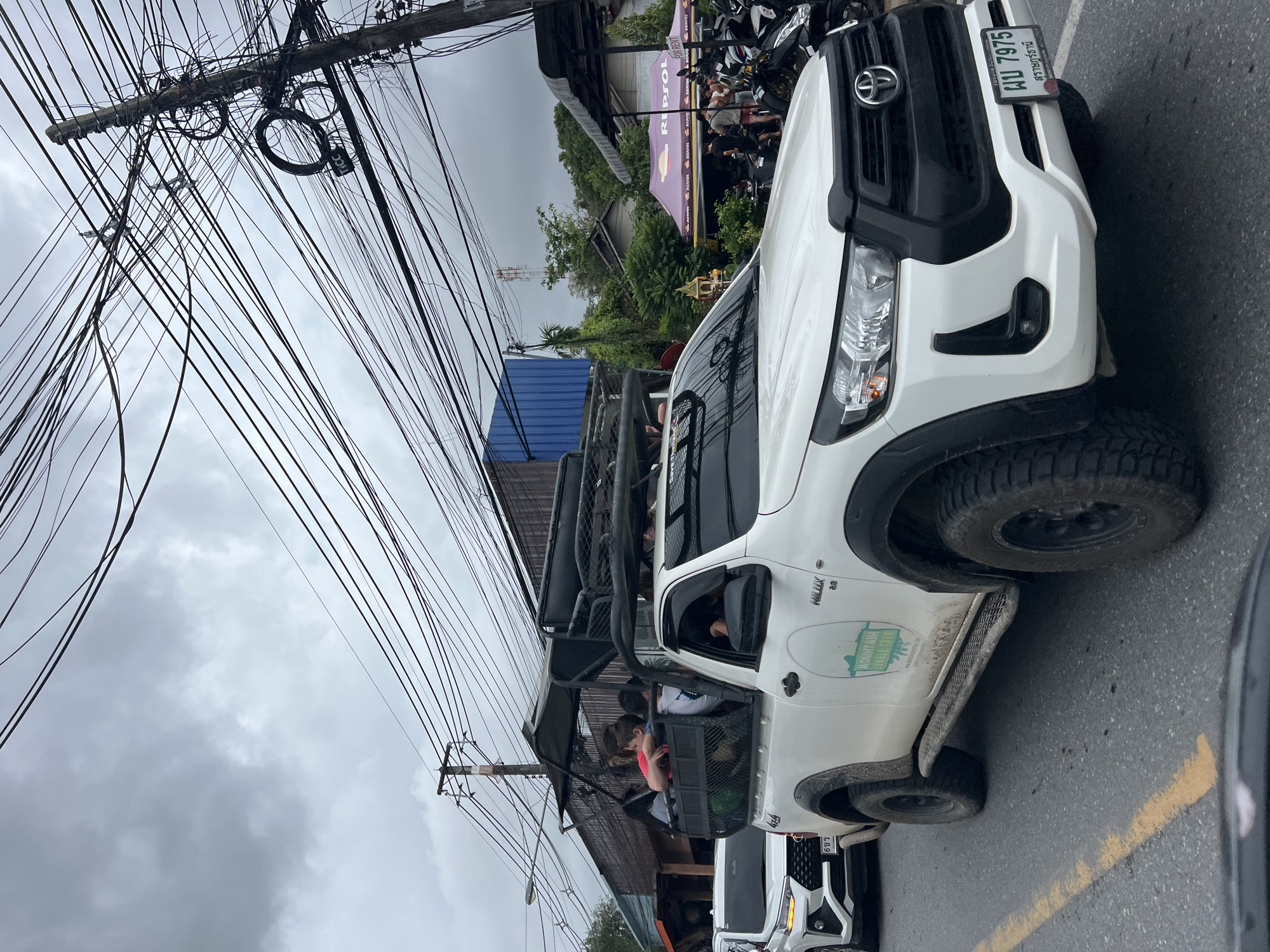 The photograph depicts a street scene in Baan Chaweng Noi, Thailand, under a cloudy sky. A white Toyota Hilux pickup truck, possibly used for a "mountain jungle tour," is prominently featured. The vehicle is occupied by several individuals in the back, within what appears to be a caged area. A second white truck is partially visible behind the first. In the background, there are buildings and parked motorcycles, including a Repsol sign. Overhead, a dense network of electrical wires obscures the sky. The license plate of the lead vehicle is blurred, but partially visible: ผบ 7975. There is a yellow lane marker painted on the asphalt road.