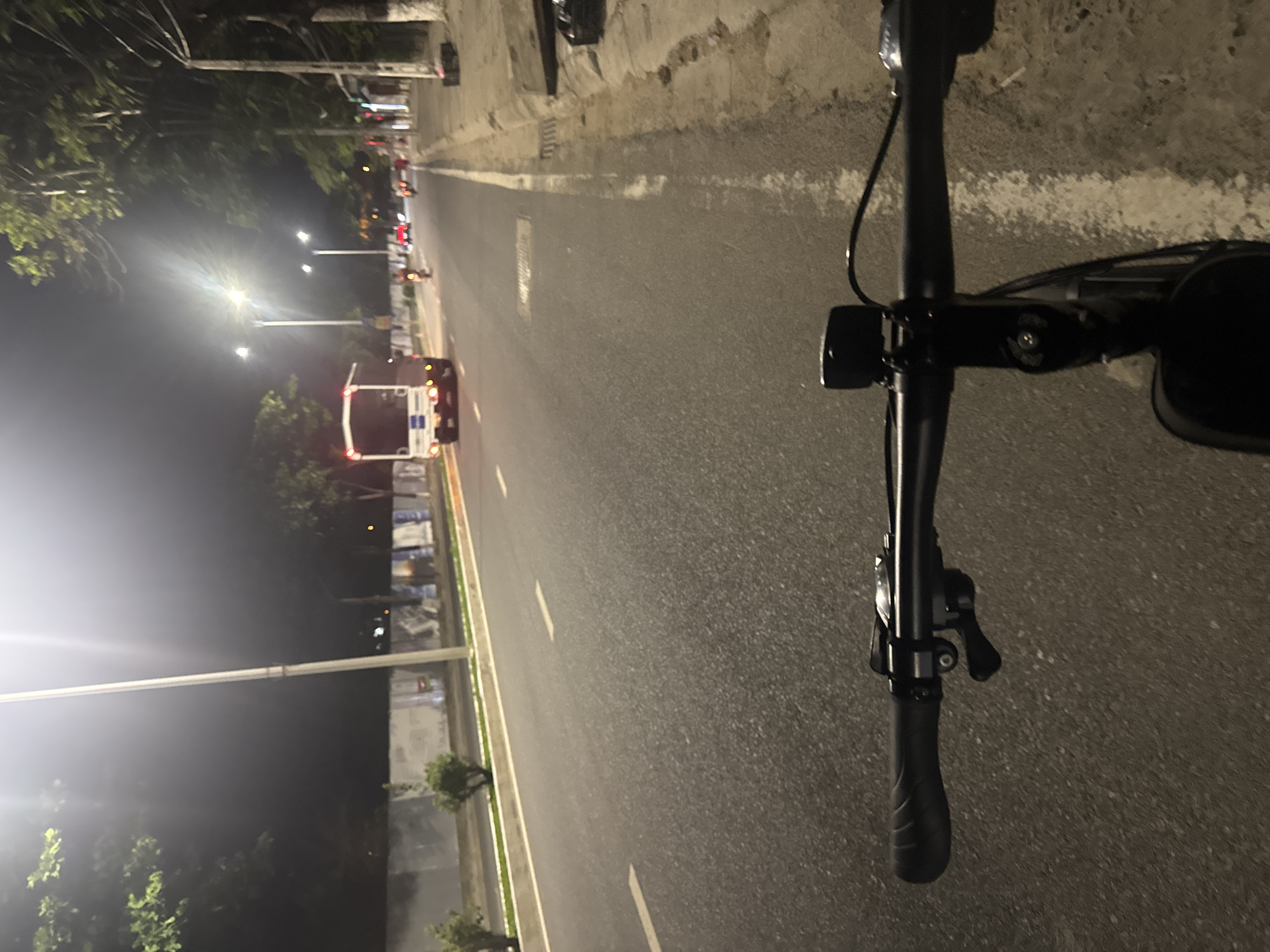 The image captures a first-person perspective from a bicycle, riding on a road at night. The road is asphalt with white lane markings, indicating a multi-lane street. To the left of the road is a low wall, and beyond that, trees and streetlights. To the right of the road is a sidewalk with some vegetation and streetlights. Several vehicles are visible on the road, including a white truck in the distance, and what appear to be motorcycles. The lighting suggests an urban or suburban environment, likely within the city of Bac Nha Trang Ward, Vietnam, given the context. The bicycle's handlebars and a mounted accessory are prominently in the foreground. The overall atmosphere is dark, illuminated by streetlights and vehicle headlights.