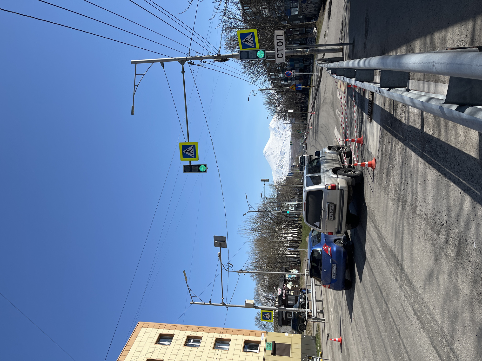 The image depicts a street scene in Petropavlovsk-Kamchatsky, Russia, on a sunny day. The view is along a street with a guardrail on the right and a multi-story building on the left. A line of vehicles is parked along the right side of the road, including a blue Suzuki and a silver SUV with license plate "X 481 OP 11" on its rear bumper. Orange traffic cones are placed along the road, particularly behind the parked cars. Overhead, a series of utility poles support wires and streetlights. Pedestrian crosswalk signs and traffic lights are also visible; all of the lights are green. In the distance, a snow-capped mountain range can be seen under a clear blue sky. Stop signs with text in Russian are visible near pedestrian crossings. The photograph is oriented in portrait format.