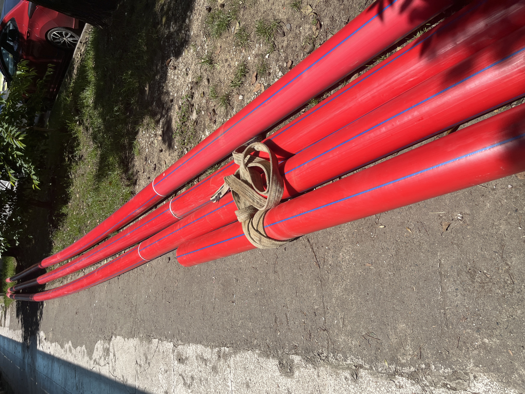 The image displays a bundle of long, bright red pipes, each marked with two parallel blue stripes along its length, resting horizontally on the ground. A tan, woven fabric strap or rope is tied around a group of these pipes, securing them together.
The pipes are laid out on a grey, paved surface, likely concrete or asphalt, which transitions into a strip of unkempt ground covered with sparse green grass, dirt, and scattered dry leaves. To the far left, a portion of a dark red car is visible, including its wheel, parked beside some green foliage. Distinct shadows, cast by unseen objects like trees or structures, fall across the pipes and the ground, indicating bright daylight and clear weather. The ends of the pipes visible on the left side of the image appear to be dark, possibly capped or a different color.
No people are present in the scene. There is no discernible text on any of the objects. The setting appears to be an outdoor area, possibly a roadside or a construction staging site, in an urban or suburban environment, consistent with the location context of Sochi, Russia. The arrangement of the pipes suggests they are materials awaiting use, possibly for underground utility work.