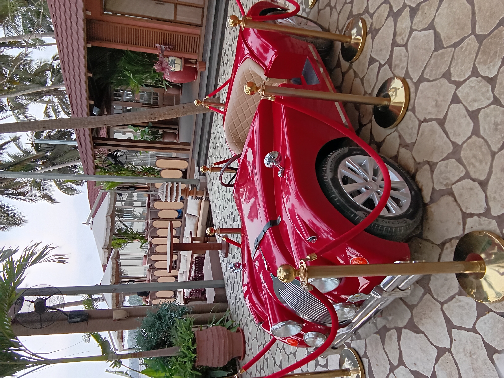 The image shows a red vintage-style car displayed outdoors. The car is roped off with red velvet ropes and gold stanchions. The surface beneath the car is paved with irregularly shaped stones.
In the background, there are glimpses of an outdoor seating area with tables and chairs, as well as palm trees. A building with a brown-tiled roof and wooden features is also visible.
Given the mention of Marna, India in the prompt, it can be inferred that the location is likely a resort, restaurant, or tourist attraction with outdoor seating and a decorative vintage car on display.