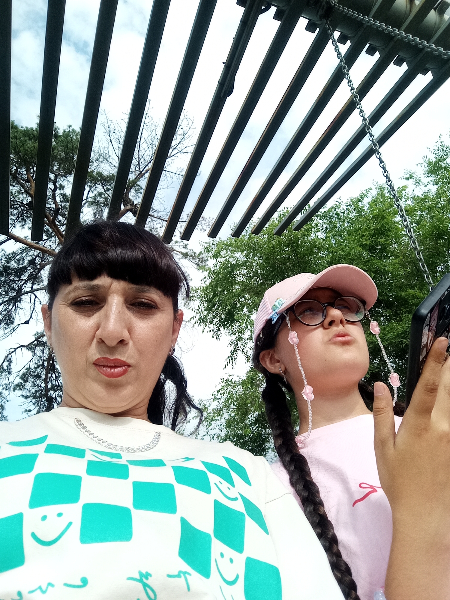 Two individuals, an adult female and a young female, are captured in a selfie outdoors. The adult female is positioned on the left of the frame, facing the camera with a neutral expression. She has dark hair with bangs and is wearing a white t-shirt adorned with a teal checkered pattern and smiley faces. A silver chain is visible around her neck. The young female is on the right, looking upwards and to her right, holding a smartphone. She is wearing a pink baseball cap, glasses, and a pink t-shirt. A pink beaded string with flower charms is attached to her glasses. Both are positioned in front of a backdrop of trees and a cloudy sky, with the upper portion of the image showing a pergola-like structure with metal bars and a chain. The overall scene suggests a casual outdoor setting, possibly a park or garden, in Belogorsk, Russia.