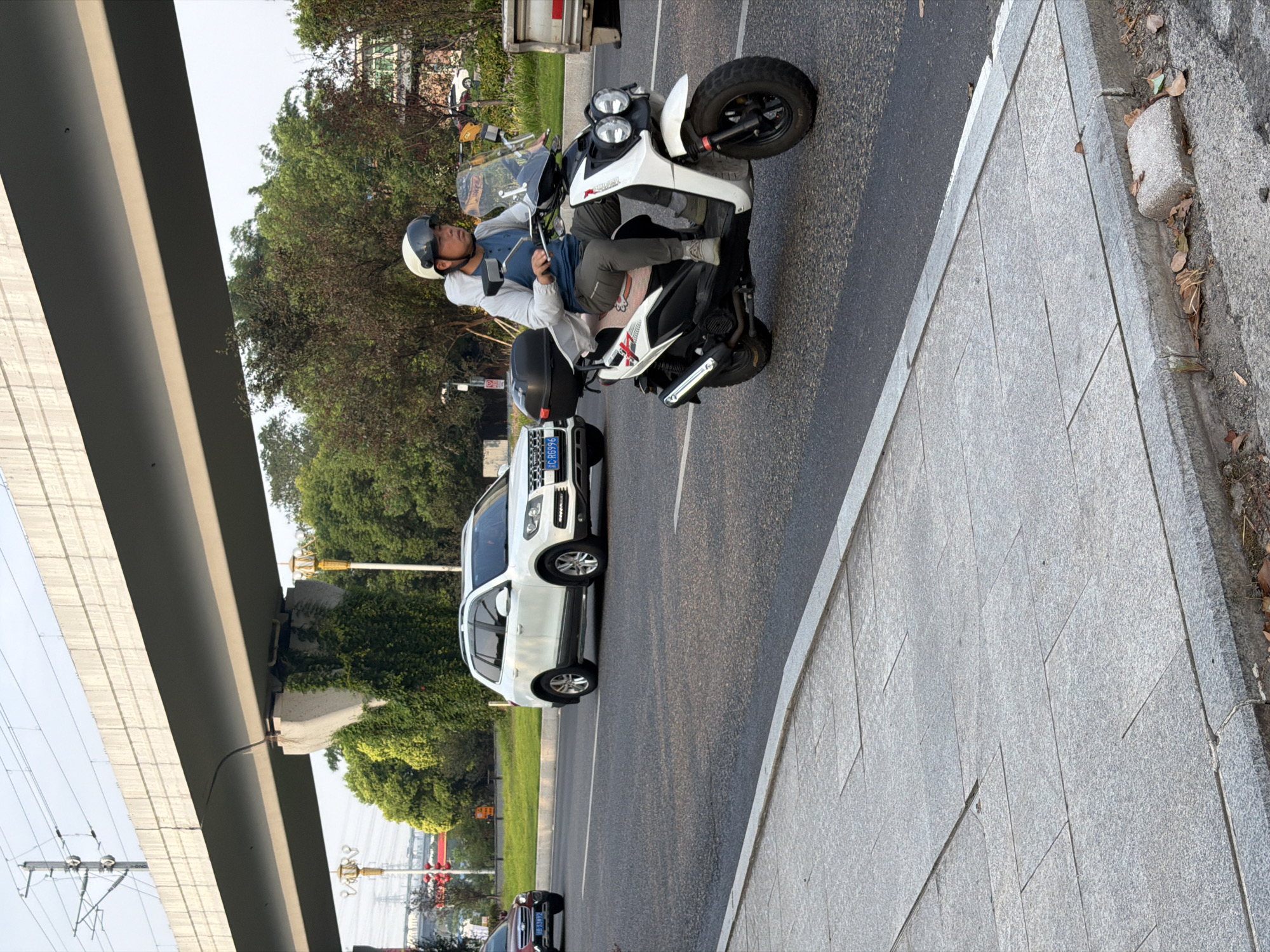In Jiangjin, China, a man wearing a white helmet and a light-colored jacket rides a white scooter on a paved road. He is in motion, his face turned slightly to the right. The scooter has two headlights and a black storage box behind the seat. To the left of the scooter, a white SUV is visible, and further left, a red car is partially visible. Overhead, a large concrete overpass supported by a concrete pillar overgrown with ivy spans the road. The sky is overcast. The road is flanked by a gray paved sidewalk with visible cracks and fallen leaves. Trees and vegetation are present on the right side of the road, and beyond them, a building with windows is discernible. The overall scene depicts urban street traffic under an elevated structure.
