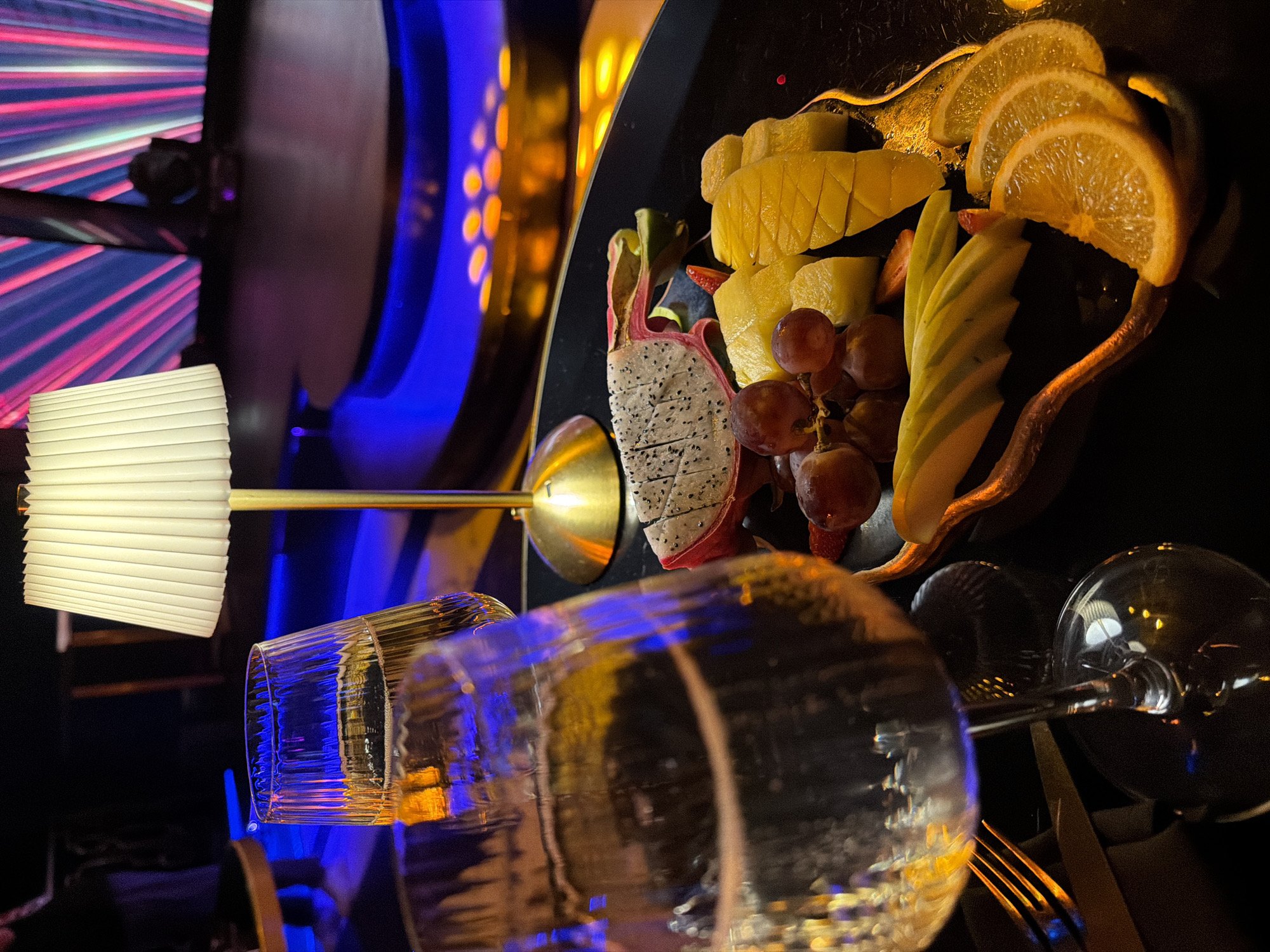 The image shows a close-up of a fruit platter and two glasses of champagne, likely in a bar or restaurant setting. The fruit platter includes sliced dragon fruit, grapes, mango, apple, and orange slices, arranged on a dark plate. One glass of champagne is in the foreground, partially obscuring a second glass behind it. A small, pleated lampshade is visible to the right of the glasses, with a gold base. The background is dark, with blurred, colorful lights, suggesting a vibrant atmosphere. The overall impression is of a luxurious or festive occasion. The image does not contain any information that would allow for identification of its specific location in Kazan, Russia.