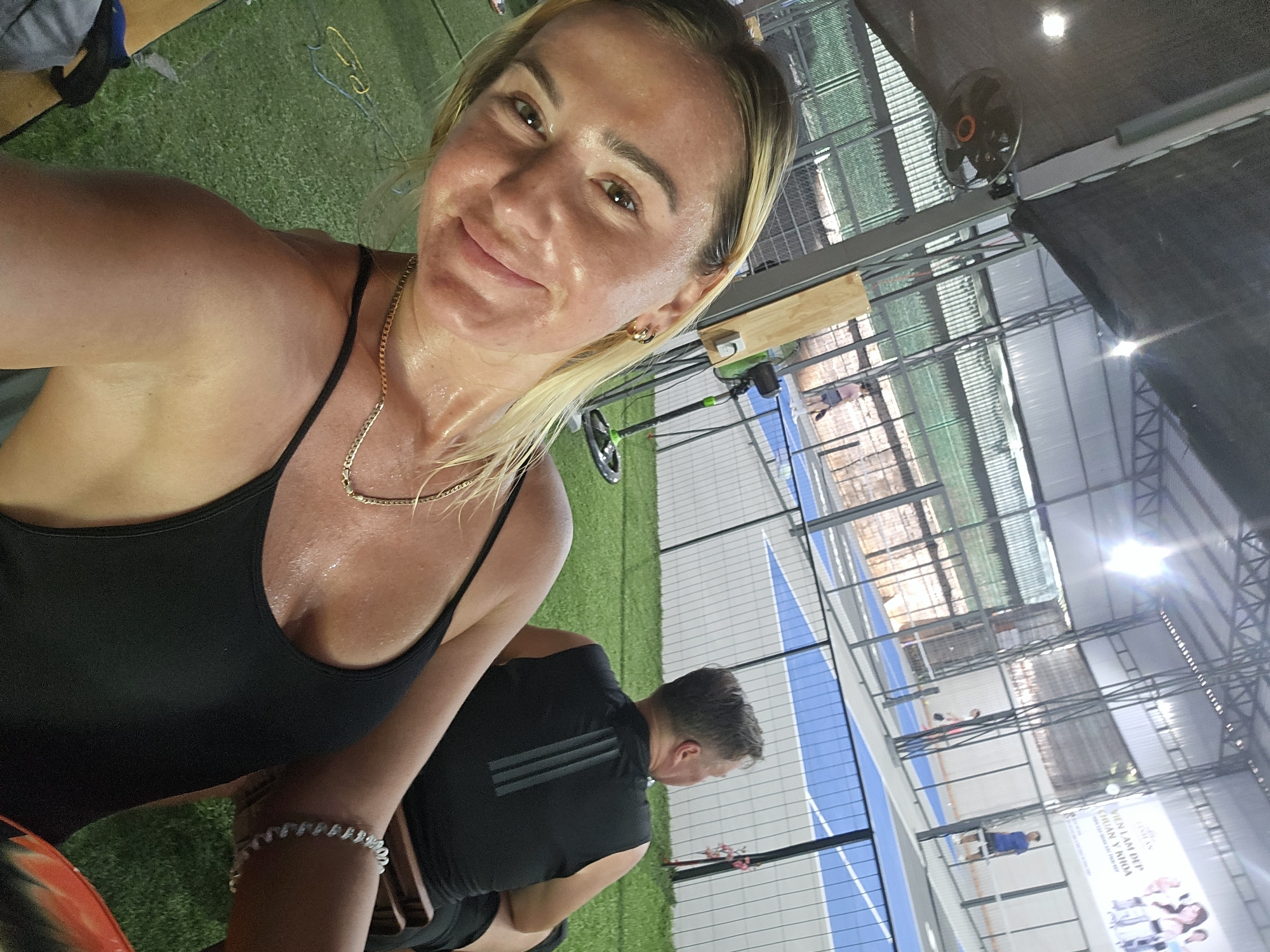 The image shows a selfie taken by a woman with blonde hair and light skin. She is wearing a black tank top and a gold necklace, and her skin appears sweaty. Her expression is a slight smile. In the background, a man in a black sleeveless shirt is visible from the back, seated on a wooden bench. The setting appears to be an indoor sports facility with a green artificial turf floor, a blue and gray court, and metal support structures for the roof. A large fan is mounted on a pole in the background. A billboard with text and images is displayed on the far wall. The location is identified as Binh Thuan Ward, Vietnam.