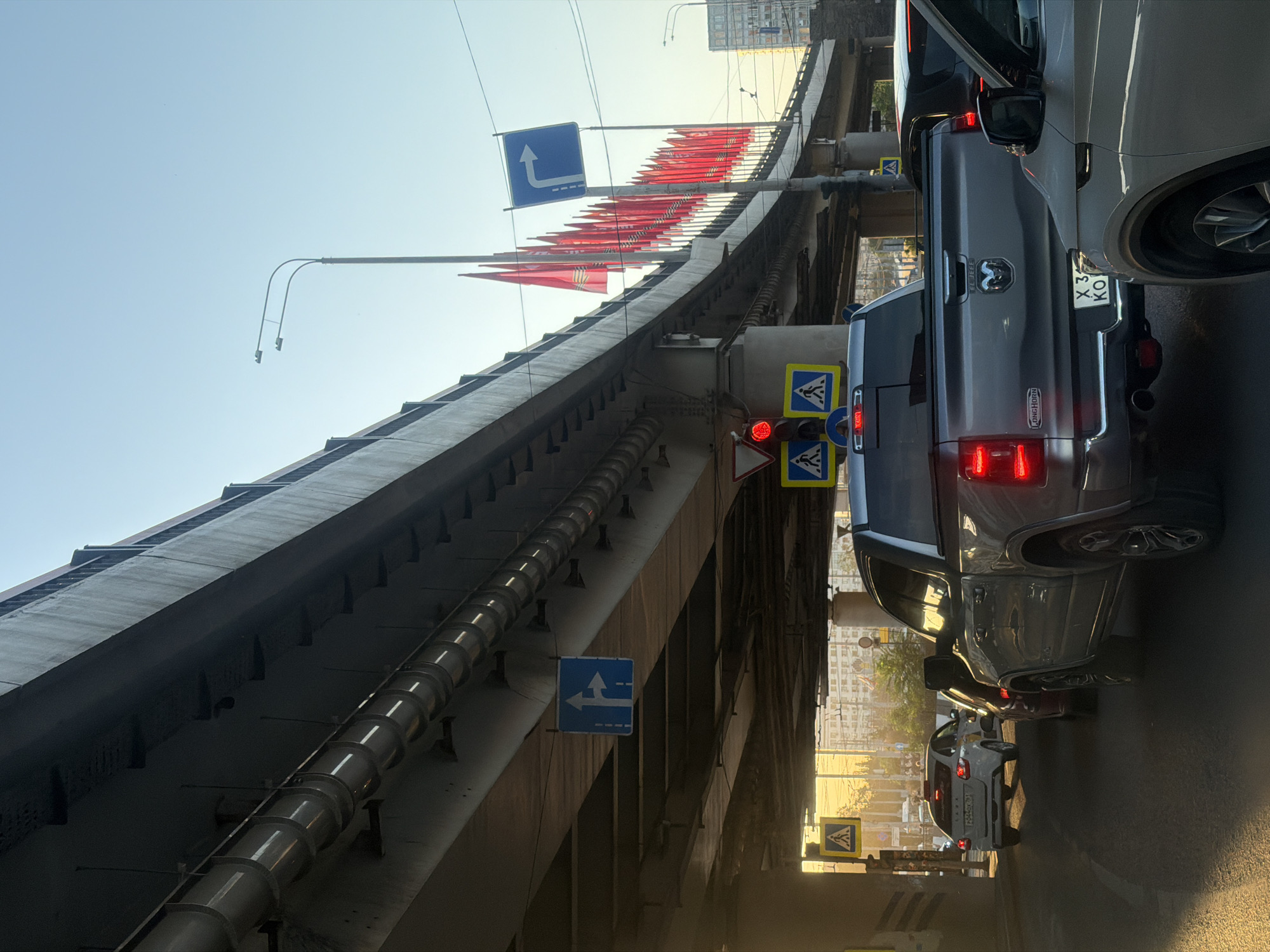 The image shows a view under a bridge in Rostov-on-Don, Russia, with heavy traffic. A line of red flags decorates the top of the bridge. Several traffic signs are visible, including pedestrian crossing and directional signs. Cars, including a grey Dodge Ram pickup truck, are stalled in traffic. The photo is taken from a low angle, emphasizing the bridge's structure and the dense urban environment. The license plate of the vehicle in front reads X347KO.