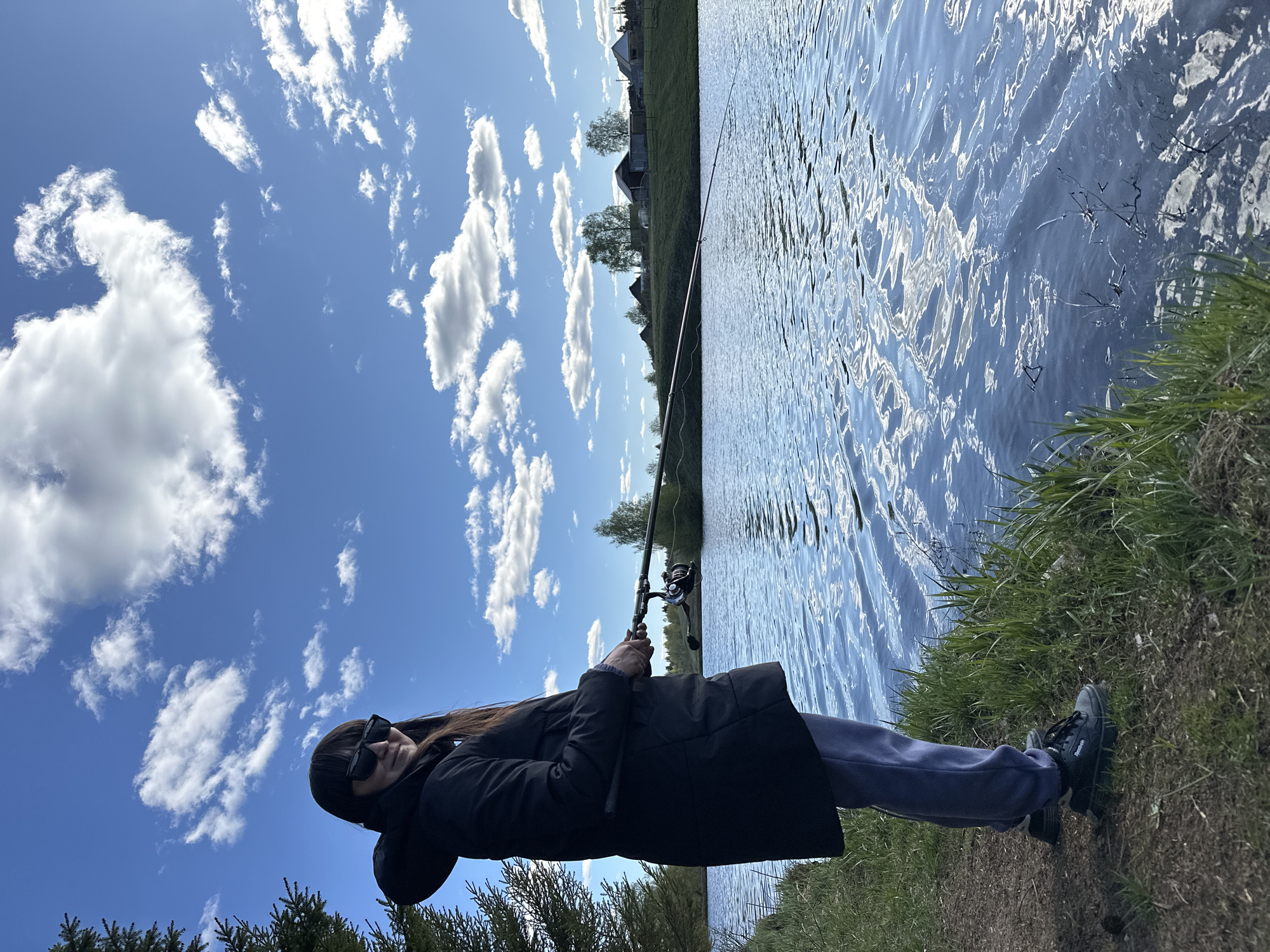 The image shows a person holding a fishing rod at the edge of a body of water, possibly a lake or river. The person is wearing a black coat, sweatpants, and sunglasses. They are standing on a grassy bank next to the water. A fishing rod extends over the water. The environment includes grass, trees, a body of water, and a sky with clouds. The location is possibly near Buranovo, Russia, based on the landscape which features rural buildings. The person appears to be fishing.