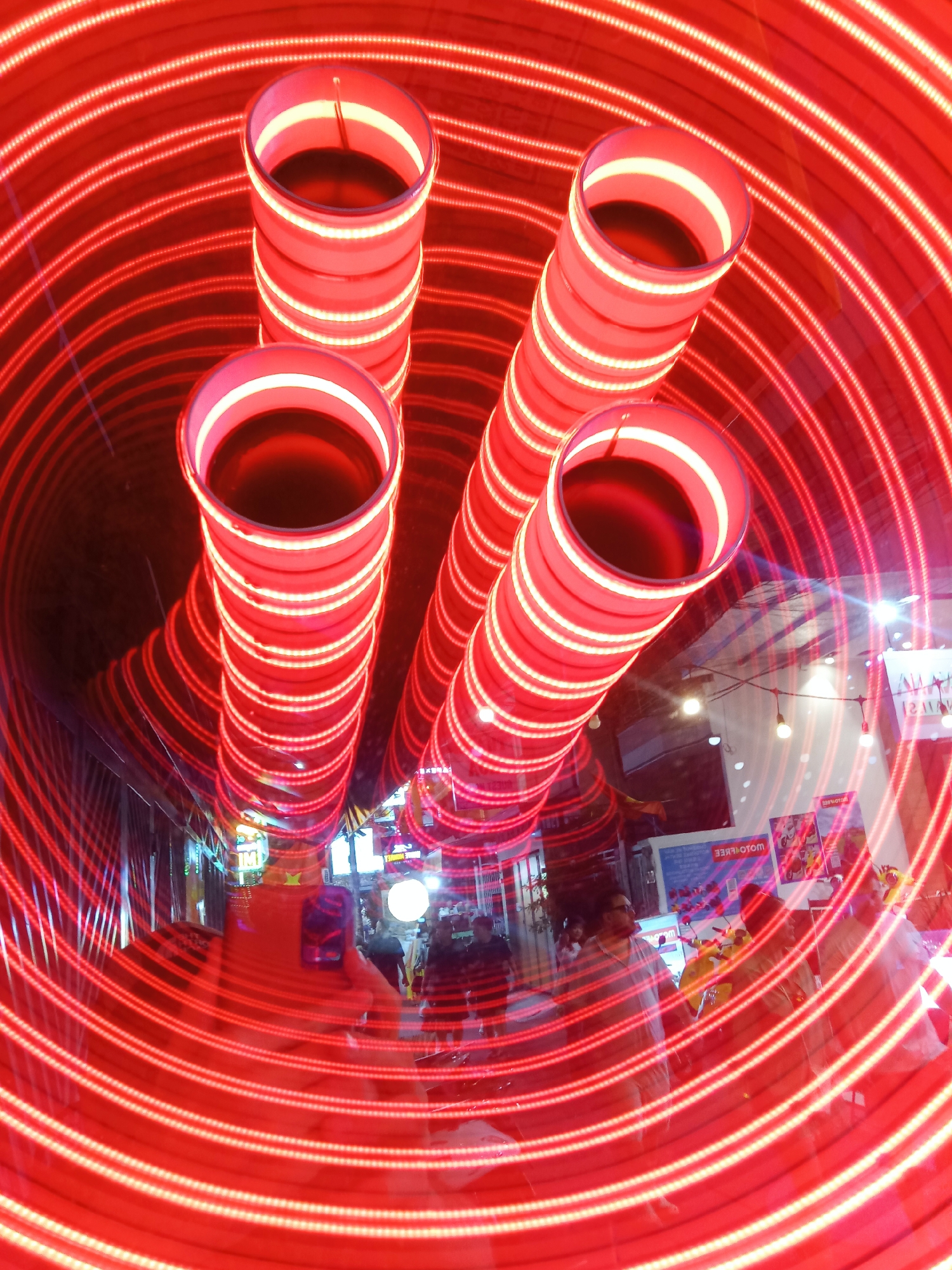 A wide-angle shot captures a crowded street in Nha Trang, Vietnam, possibly during an event or festival. The most striking feature is the red neon light installation, comprising several cylindrical shapes arranged vertically, surrounded by concentric circles of red lights creating a tunnel effect. The scene is taken from within the light installation, with people visible in the background, some appearing blurred due to the focus on the lights. Commercial storefronts and signage are visible in the distance, indicating a busy commercial area. Several individuals can be identified, some taking pictures of the installation. A yellow scooter is visible to the right of the crowd. A blue and white sign with mirrored text hangs on the wall on the right.