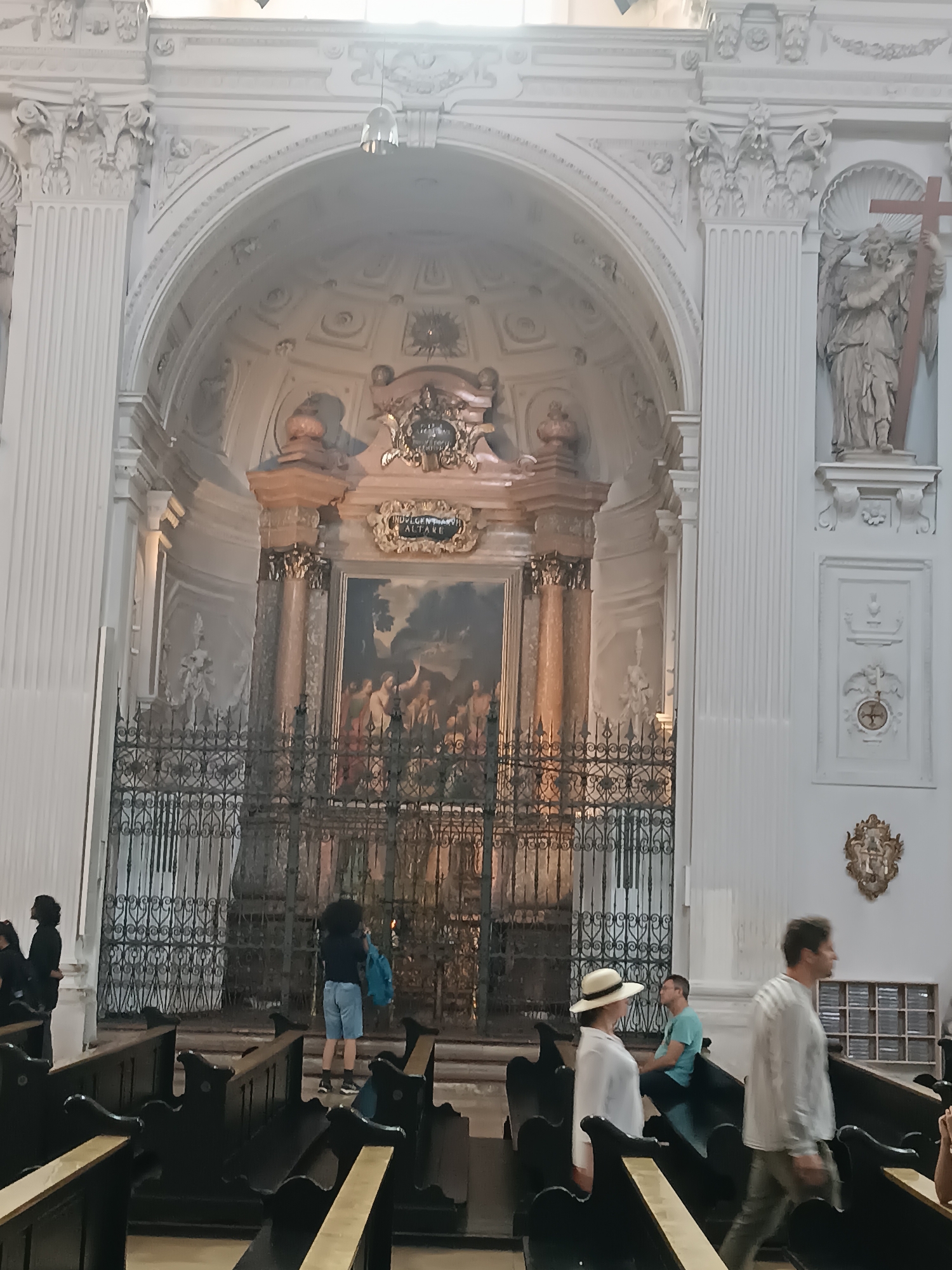The image captures an interior view of a grand, ornate church, likely the Hofkirche in Munich, Germany. The scene is dominated by classical architecture with white columns, intricate carvings, and a high arched ceiling. In the central area, a decorative iron grate separates the nave from the altar, behind which a large painting is displayed. Several people are present in the church. In the foreground, dark wooden pews are arranged in rows. To the left, a woman in a white outfit and a hat is seated, facing away from the camera. To her right, a man in a light-colored striped shirt is walking away. Further back, near the altar, a person is standing with a blue bag. Another person in shorts and a blue shirt is seated on a pew. On the right side of the image, an angelic sculpture holding a cross is visible on a pedestal, and further down, a man in a light-colored shirt is walking away from the camera. The lighting is soft, suggesting ambient light filtering from above and possibly some artificial lighting within the church.