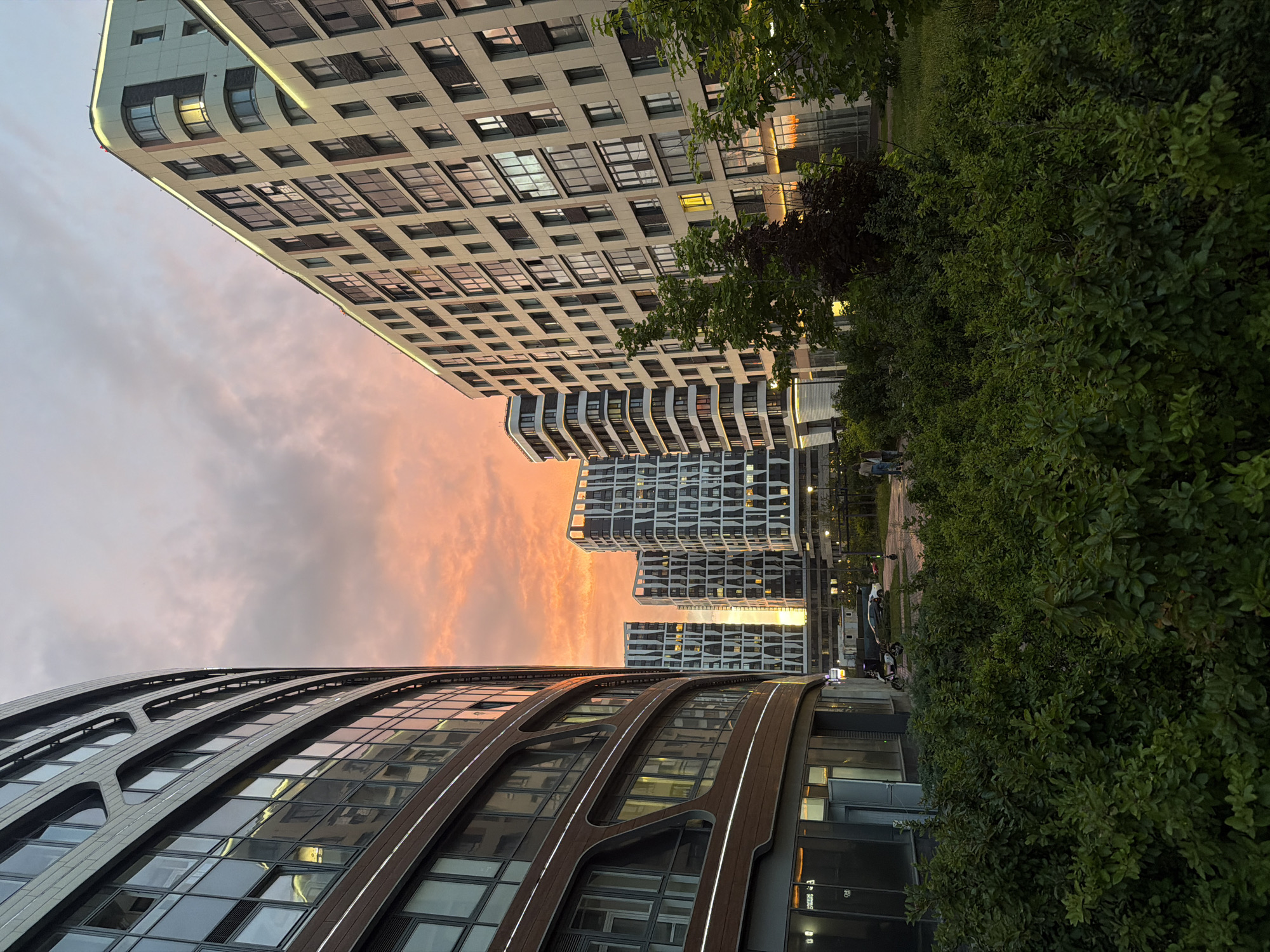 This is a wide-angle, eye-level shot of modern apartment buildings at dusk in Moscow, Russia, likely in the northeastern part of the city. The sky is filled with dramatic sunset clouds in shades of orange, pink, and purple. In the foreground, a dense, well-maintained hedge of green bushes lines the bottom of the frame. Behind the hedge, a paved area shows a parked scooter and two cars. Two individuals are walking away from the viewer on this paved area. The architecture is contemporary, with large glass windows and geometric facades. The building on the left is curved and features brown and silver elements. To its right are several taller, rectangular buildings with white and dark grey patterned facades. Lights are visible inside some of the apartments. The scene suggests a peaceful evening in a developed urban residential area.