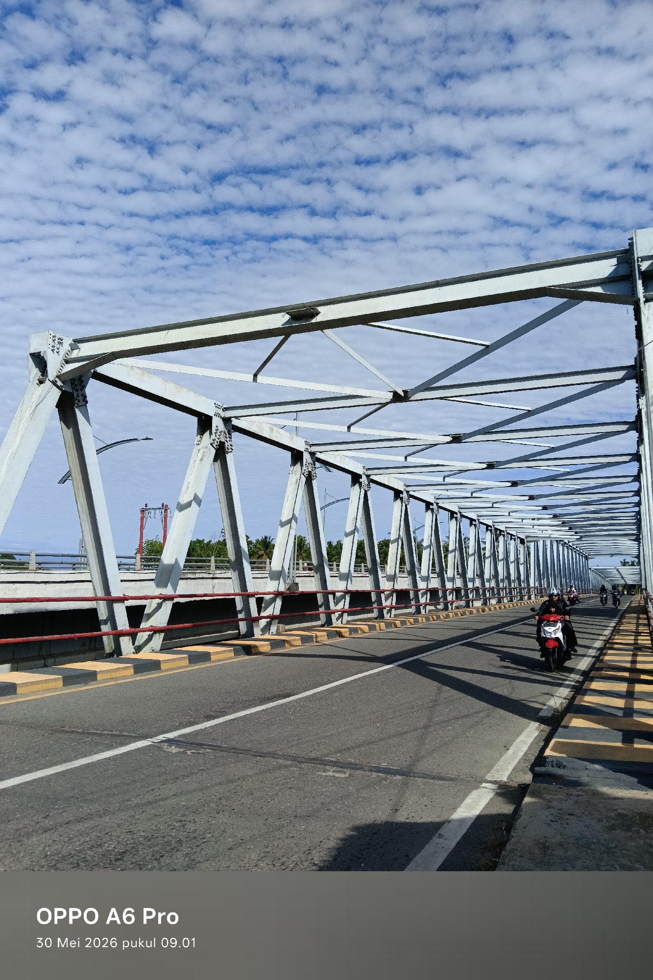 The image shows a steel truss bridge in Neuboknaleung, Indonesia. A line of motorcycles are on the bridge. The bridge has red railings and yellow-and-black striped borders. The sky is light blue with scattered white clouds. The horizon shows trees on either side of the river.