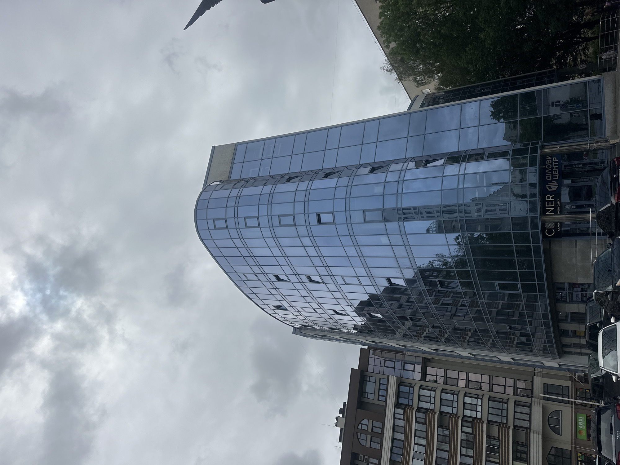 The image captures a modern, curved glass-fronted office building in the city of Ternopil, Ukraine. The building is characterized by its reflective blue glass facade, which shows reflections of other buildings and the cloudy sky. Adjacent to it is another apartment building with a more traditional architectural style, featuring balconies and a patterned facade. The scene is set on a cloudy day, with the sky overcast and diffused light. In the foreground, several dark-colored cars are parked, suggesting a street-level view. The lower section of the glass building displays signage with Ukrainian text, indicating it is a business center. To the right of the office building, a dense green tree is visible, adding a touch of nature to the urban landscape. A portion of a dark, pointed structure, possibly a sculpture or architectural element, is visible in the upper right corner of the image.