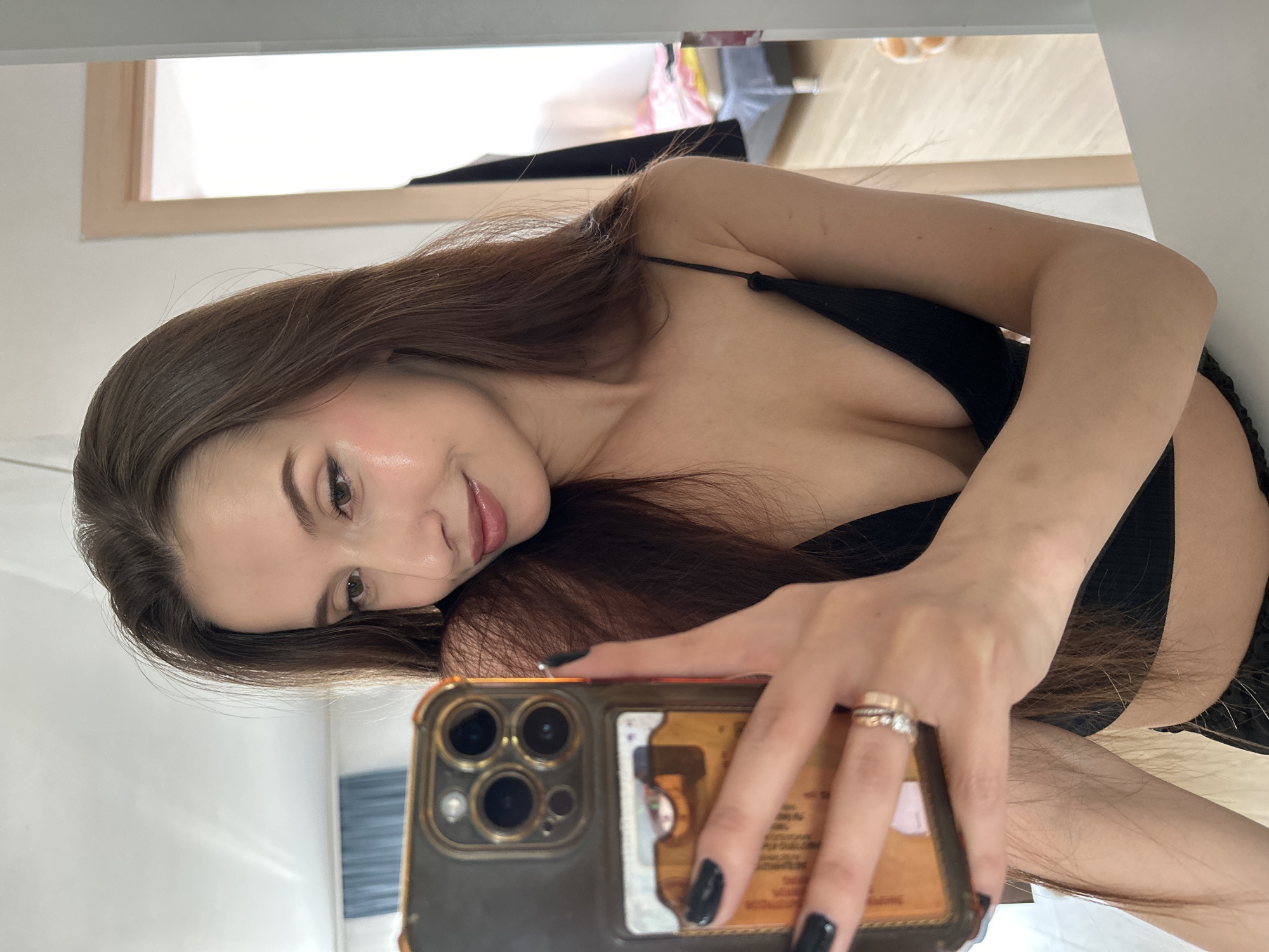 A woman with long brown hair takes a selfie. She is wearing a black top and holding a gold-colored iPhone. Her nails are painted black, and she has two rings on her finger. The background shows a white wall with a framed window and some indistinct objects. It is not possible to discern the location in Yekaterinburg, Russia, from the image.
