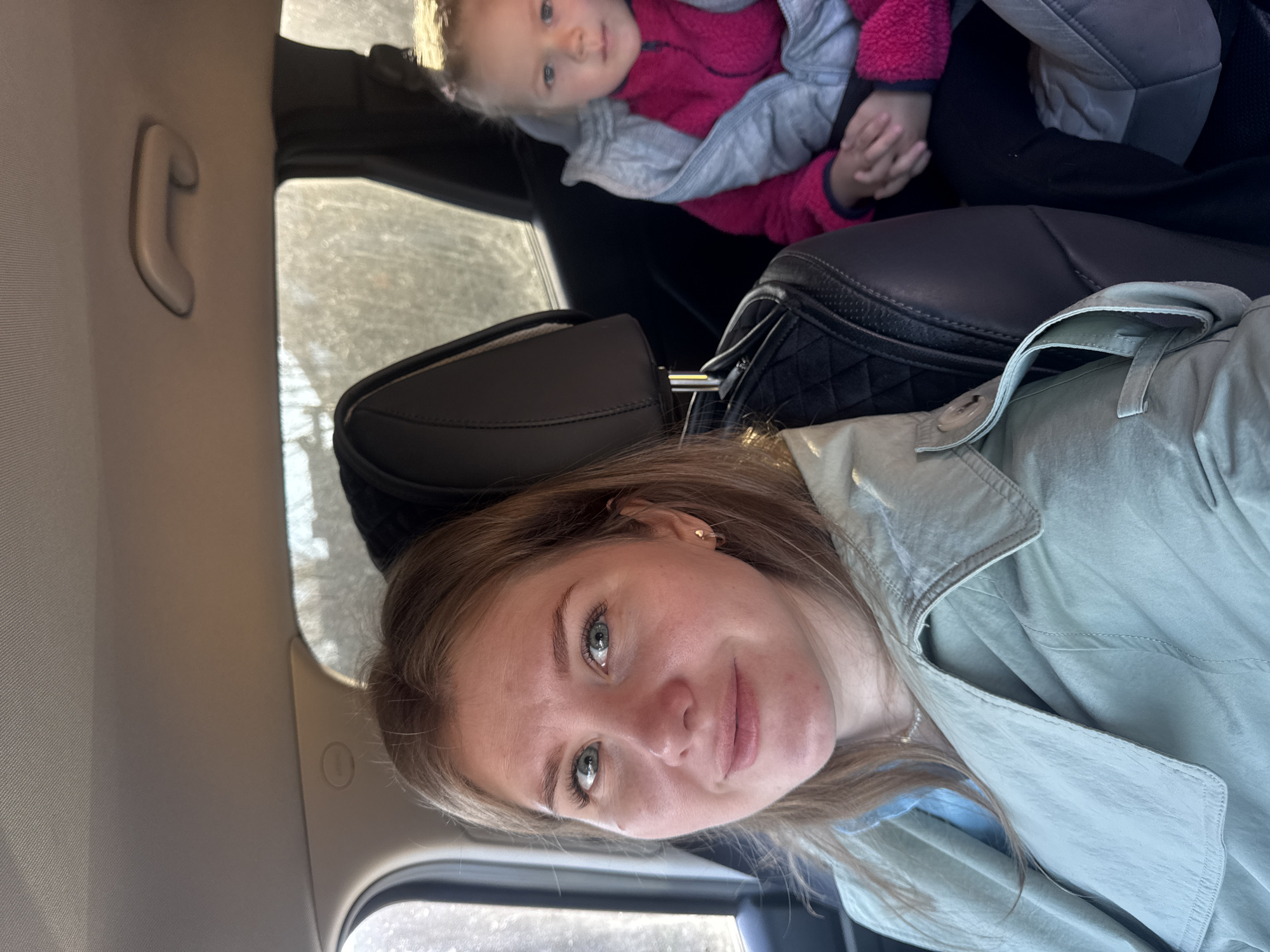 The image depicts a young woman in the foreground and a child in the background, both inside a vehicle, likely captured as a selfie. The young woman has light brown hair, light-colored eyes (appearing greenish-blue), and a subtle smile. She is wearing a light greenish-gray trench coat or jacket over a lighter blue top. A delicate silver necklace is visible on her neck.
Behind the woman, a young child, possibly a toddler, is seated in a car seat. The child has blonde hair and light-colored eyes, looking towards the camera with a neutral expression. They are dressed in a bright pink fleece top and a grey hooded jacket or coat. Part of the car seat's headrest and side cushioning is visible.
The interior of the car features a dark leather or synthetic headrest for the woman's seat. The car's ceiling and pillars are light beige, with a grey overhead grab handle visible on the left side of the frame. Daylight illuminates the interior, suggesting it is daytime. The surroundings outside the car windows are bright but indistinct. There is a glimpse of another dark bag or object next to the woman's seat, and part of a quilted black bag near the child. No visible text is present in the image. The setting is a car, potentially in Горный, Russia.