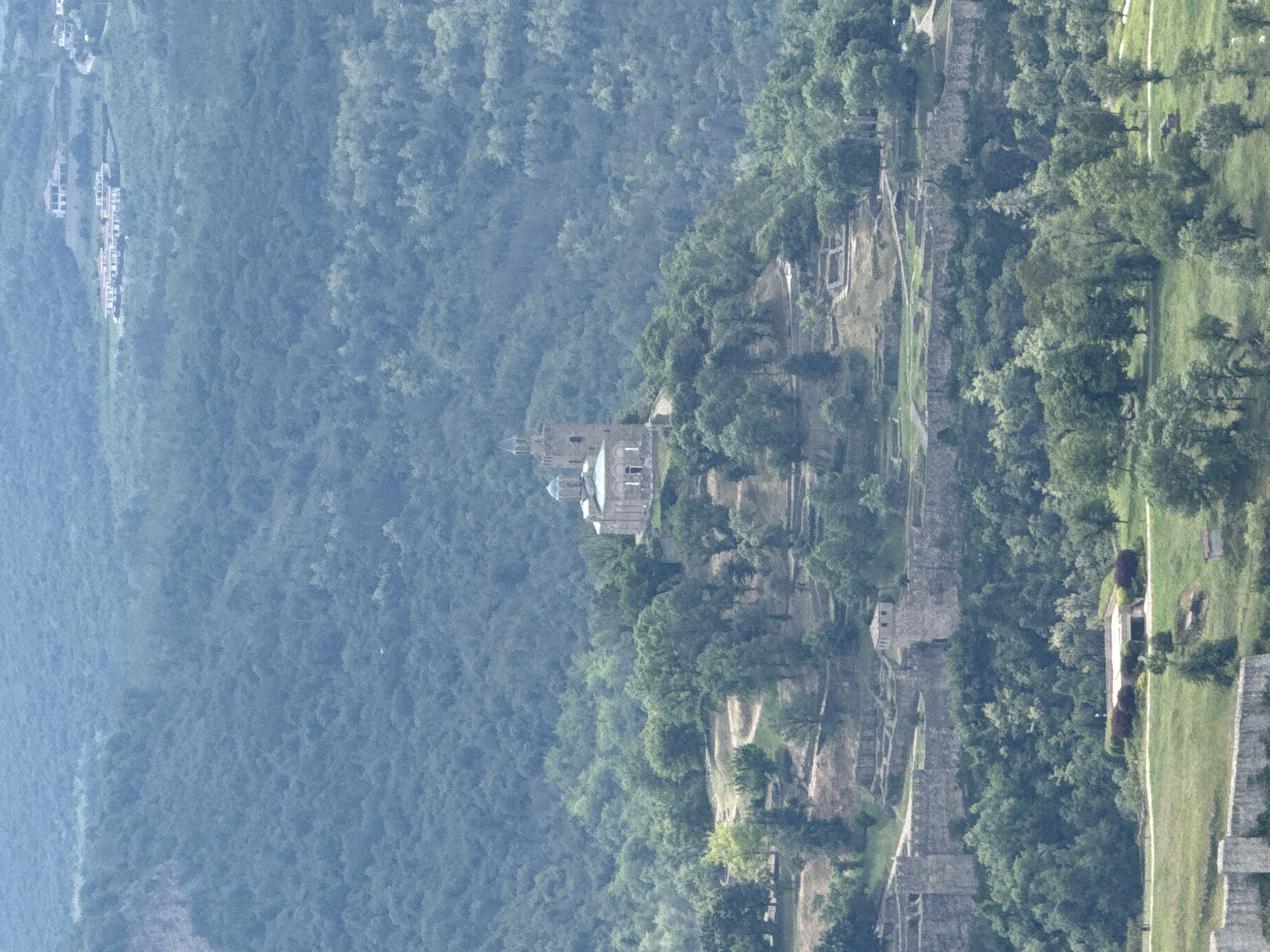 This image shows a section of the Tsarevets Fortress in Veliko Tarnovo, Bulgaria, positioned atop a forested hill. The architecture is predominantly stone ruins, interspersed with trees and greenery. The eastern part of the fortress is visible and a part of the city is observed in the background. The landscape consists of dense forests covering the surrounding hills. The sky is obscured, indicating an overcast day.