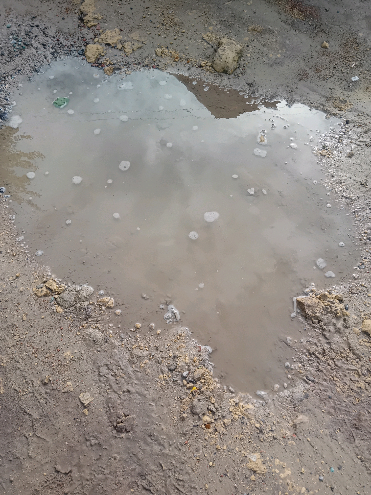 A muddy puddle is situated on a dirt or gravel surface. The water is cloudy and reflects the sky. Several small bubbles are visible on the surface of the water. The edges of the puddle are surrounded by damp earth and small rocks and pebbles. There is a green object, possibly a piece of plastic debris, partially submerged in the water on the left side of the puddle. The environment appears to be outdoors, possibly a rural road or unpaved area.