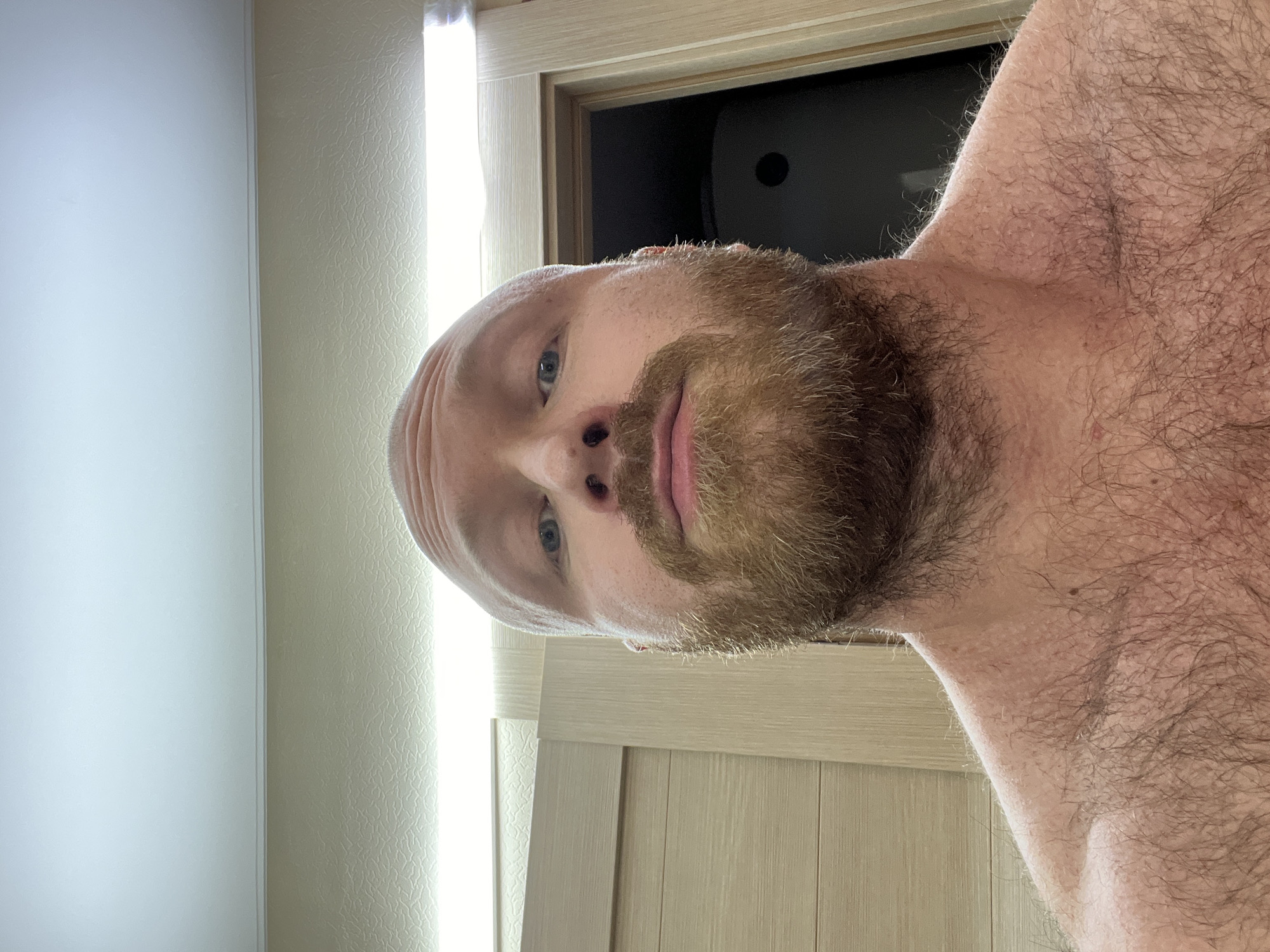 A close-up, low-angle selfie of a bald, fair-skinned man with a full, reddish-brown beard. He has blue eyes. He is shirtless. The background includes a white wall with a vertical light fixture, a light-colored wood-paneled door, and a glimpse into a dark room with a black object on the wall. There is no information suggesting this image was taken in Noviy, Russia.